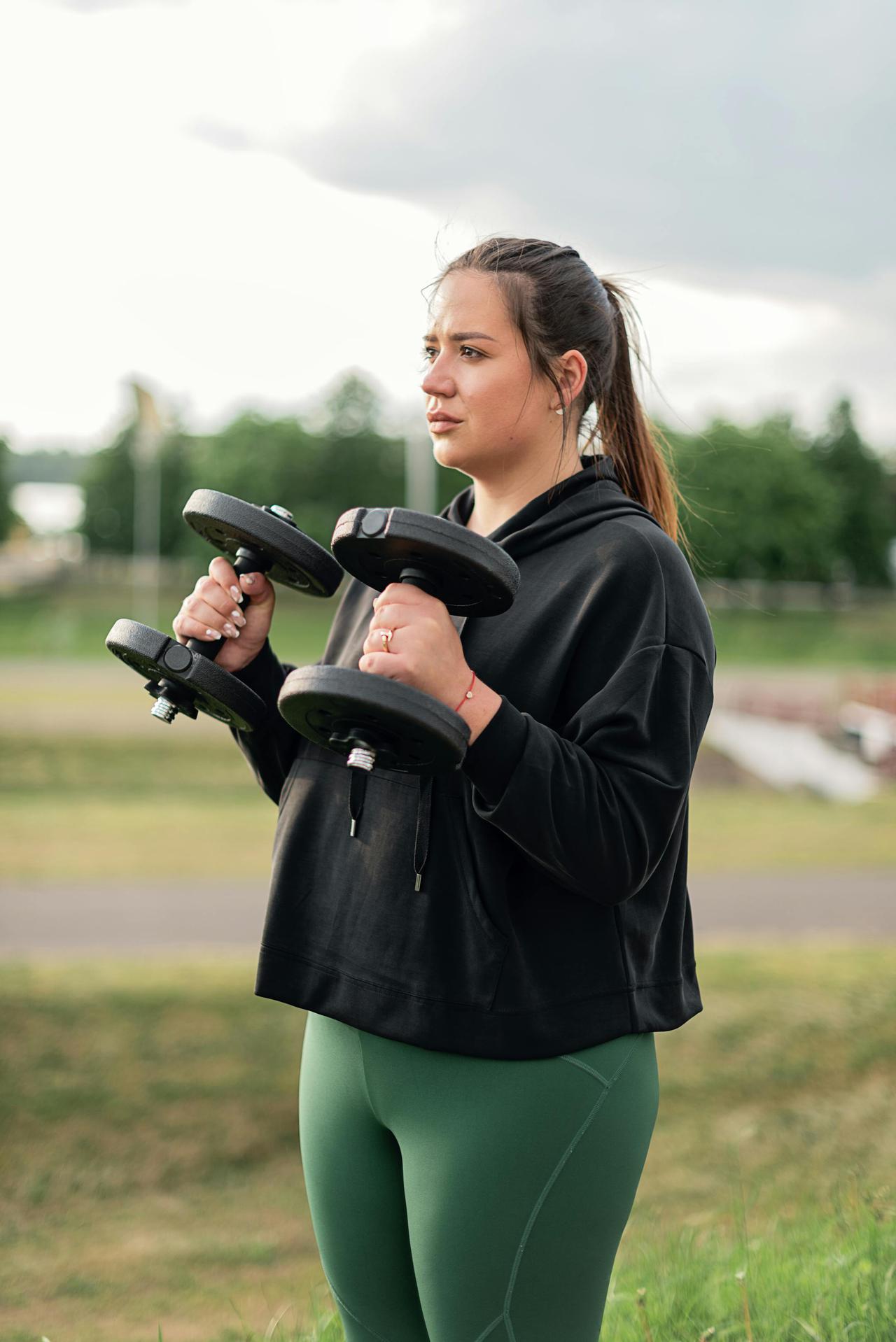 Woman in active wear lifting dumbbells outdoors, promoting fitness and wellness.
