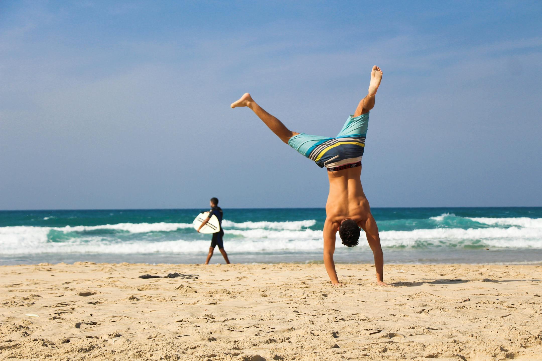 Man doing a handstand on a sunny beach with surfer in background, evoking a sense of freedom and joy.