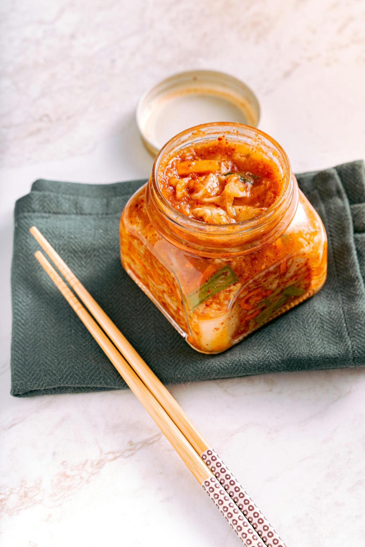 A jar of traditional homemade kimchi on a marble surface with wooden chopsticks beside it.