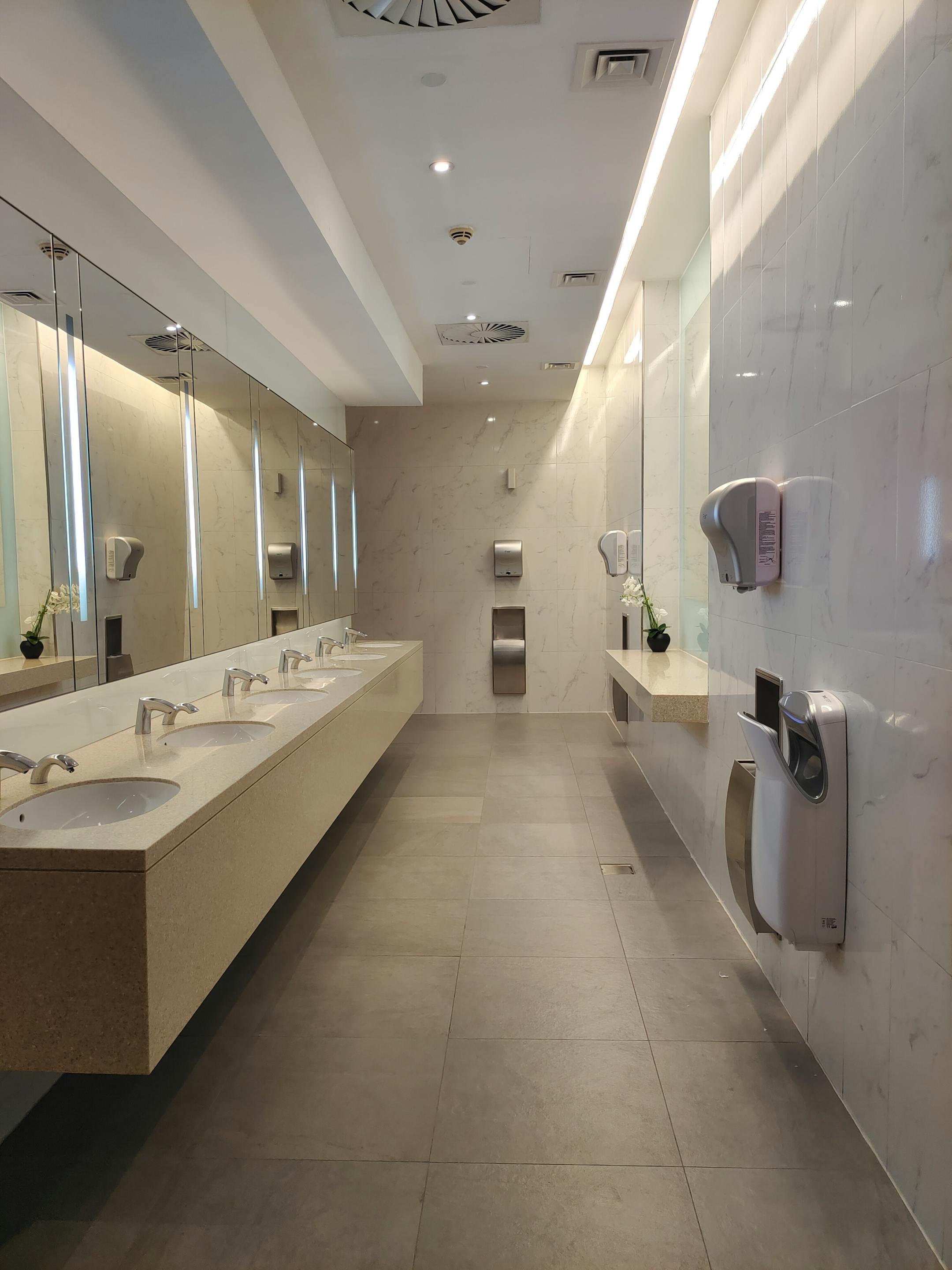 Spacious, well-lit modern public restroom with marble and beige tones, featuring multiple sinks and hand dryers.