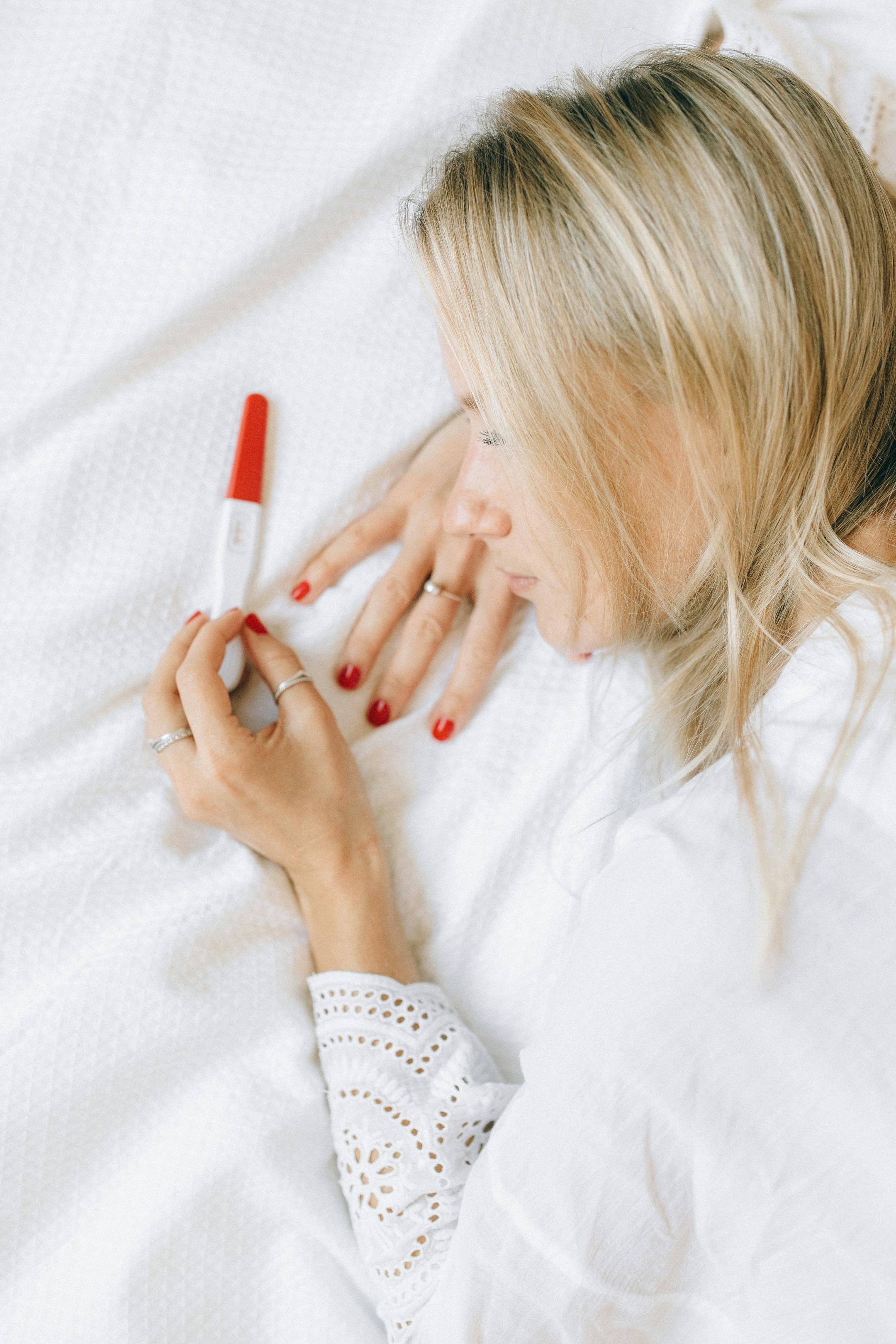A woman lying on a bed looking at a positive pregnancy test with a joyful expression.