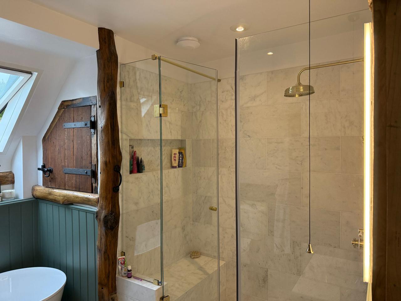 Luxury marble wetroom with brushed brass shower fittings, oak beam feature, and walk-in glass enclosure — Tiled by Coventry Tiling.