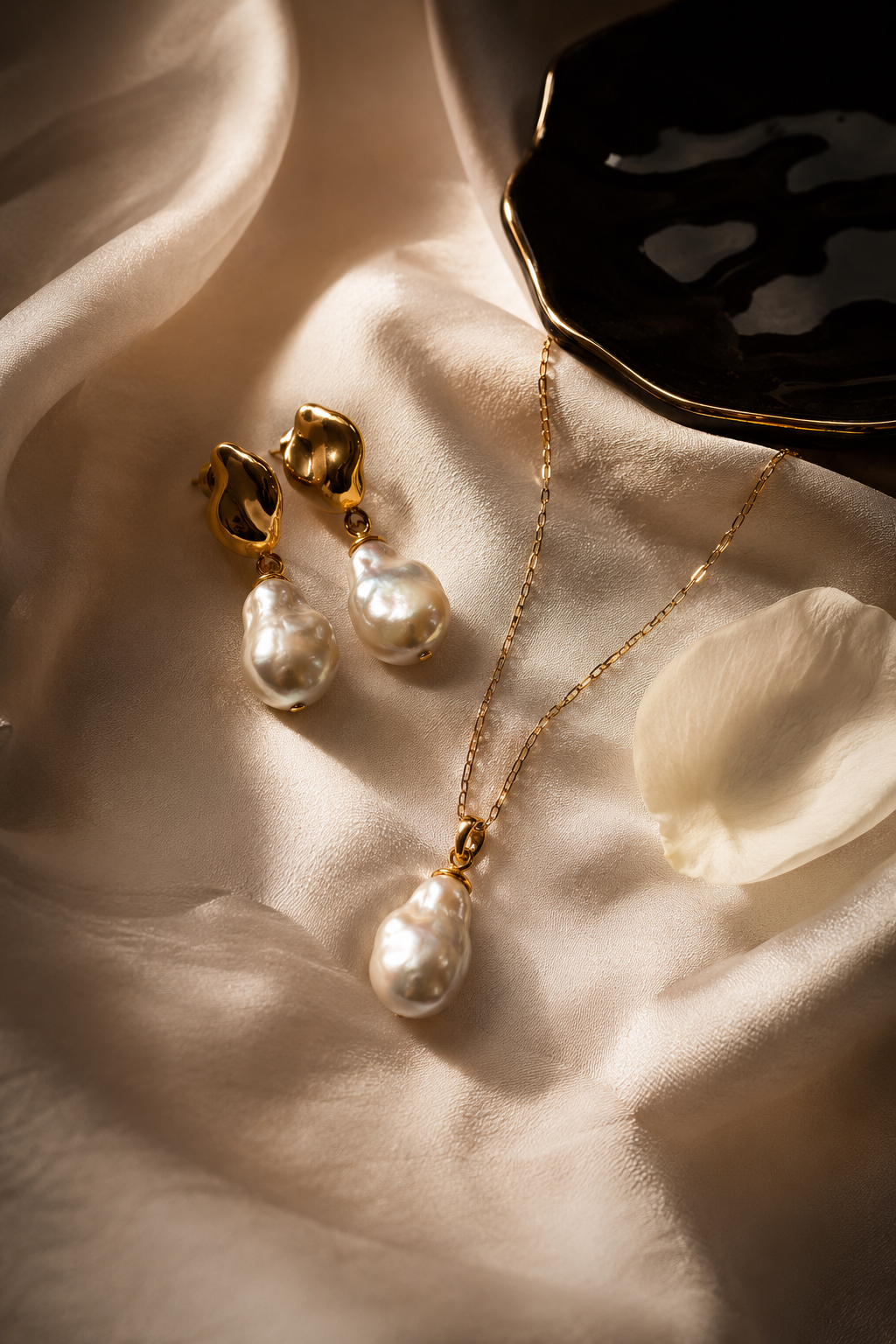 Flat-lay of baroque pearl earrings and gold pendant necklace on cream silk with a black lacquer dish and rose