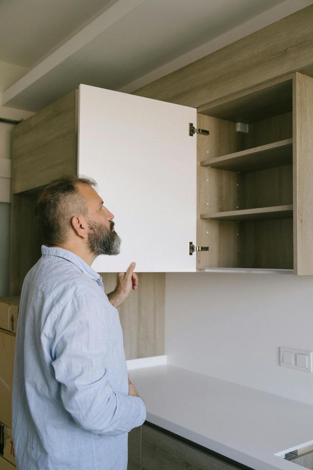 Bearded man in a blue shirt checking a kitchen cabinet in a modern indoor setting.