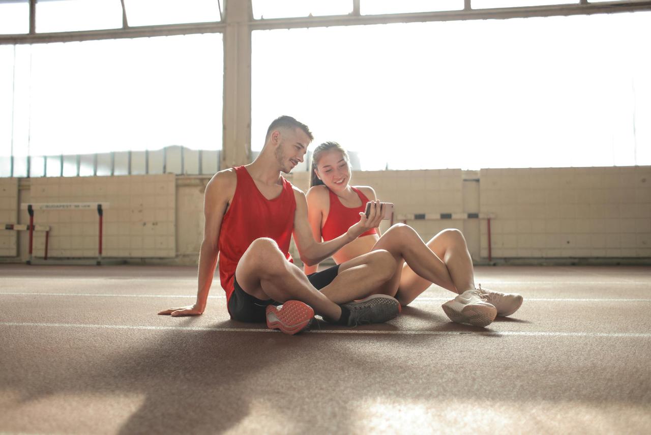 Happy young sportsmen sitting with crossed legs on floor and surfing smartphone together at sports hall