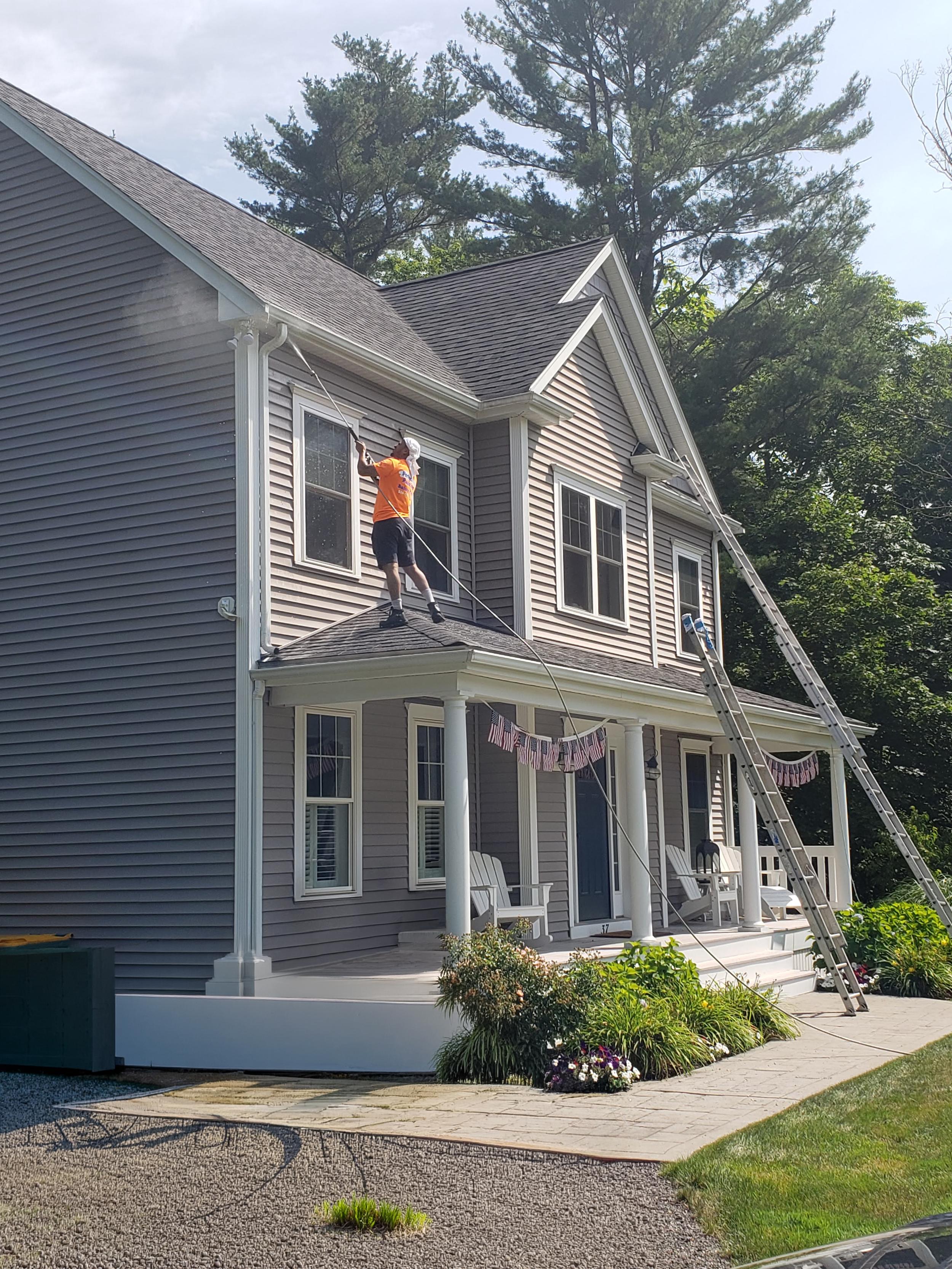 male-on-ladder-painting-exterior-of-grey-house-with-white-trim-surrounded-by-trees-shrubs-and-flowers