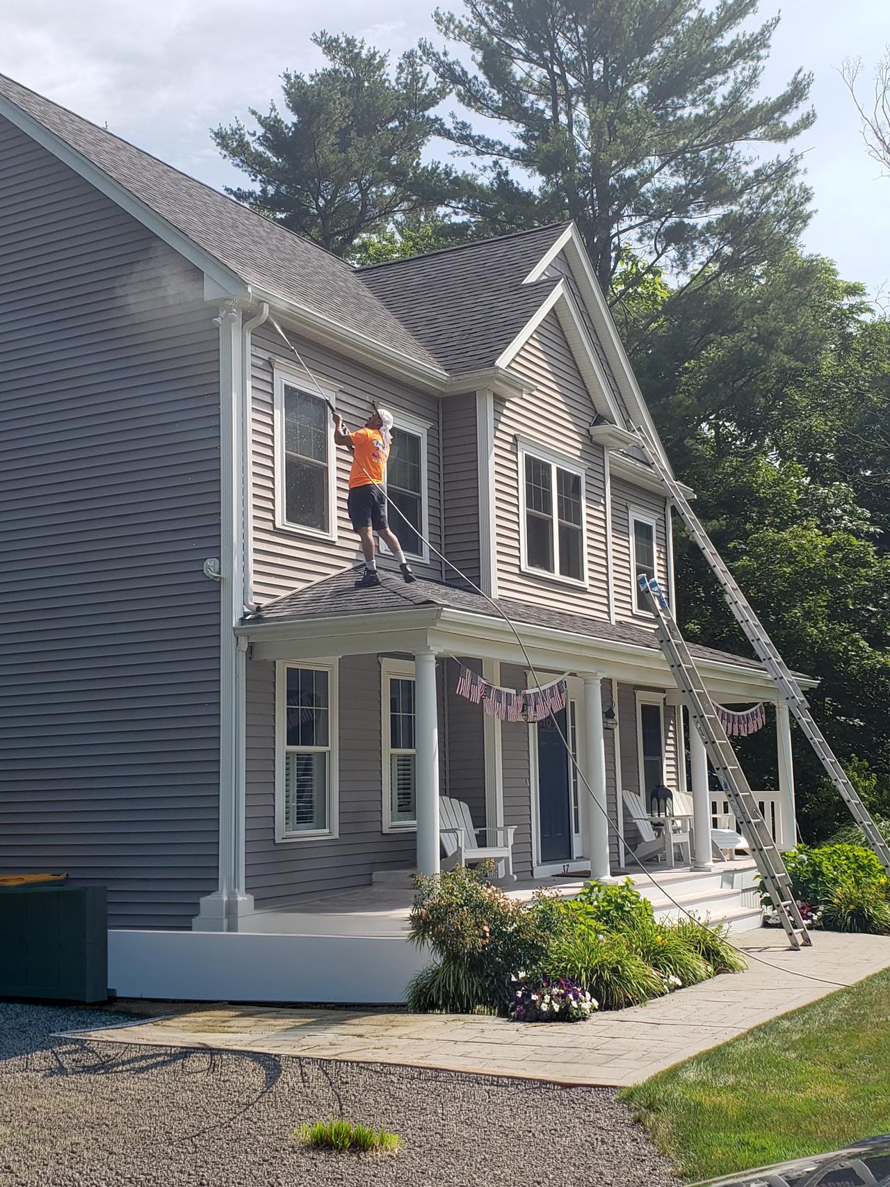 male-on-ladder-painting-exterior-of-grey-house-with-white-trim-surrounded-by-trees-shrubs-and-flowers