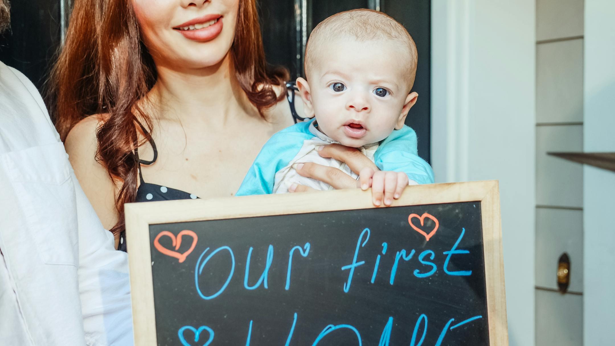 Happy family holding chalkboard sign announcing 'Our first home' with baby in the doorway.