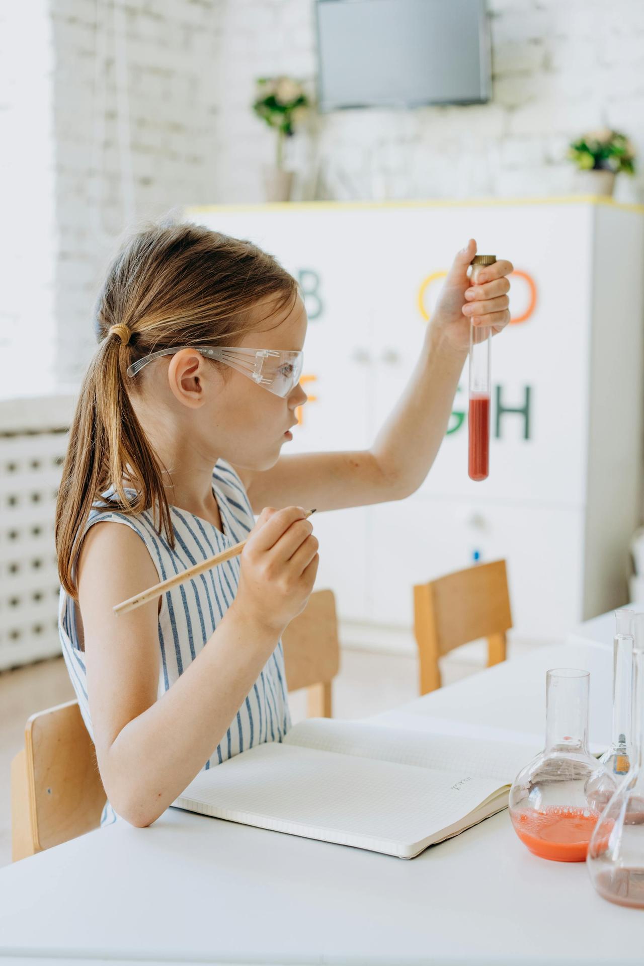 A young girl wearing safety glasses performs a chemistry experiment using test tubes in a classroom.