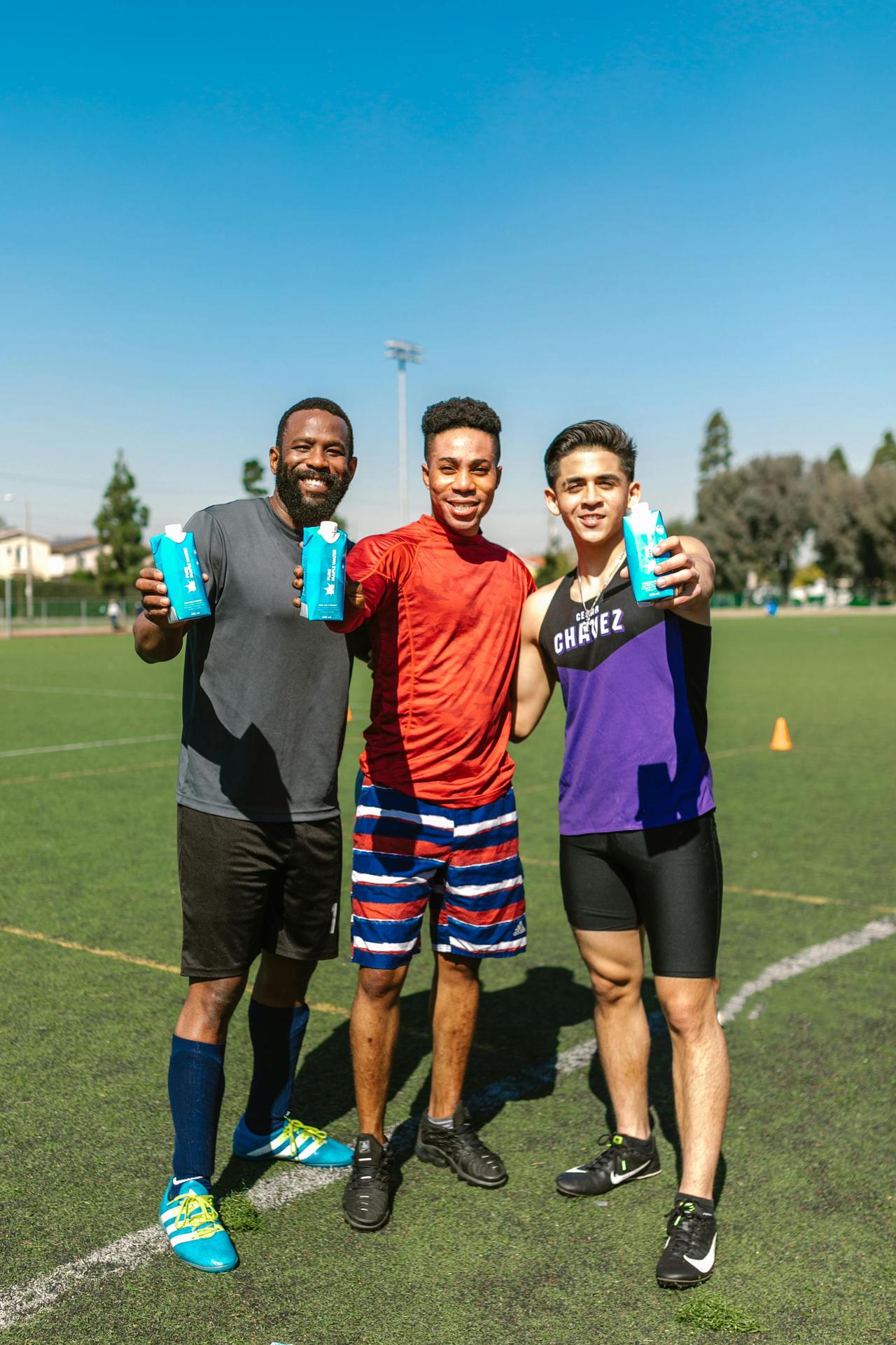 Three friends smiling and holding drinks on a sports field under a clear blue sky.