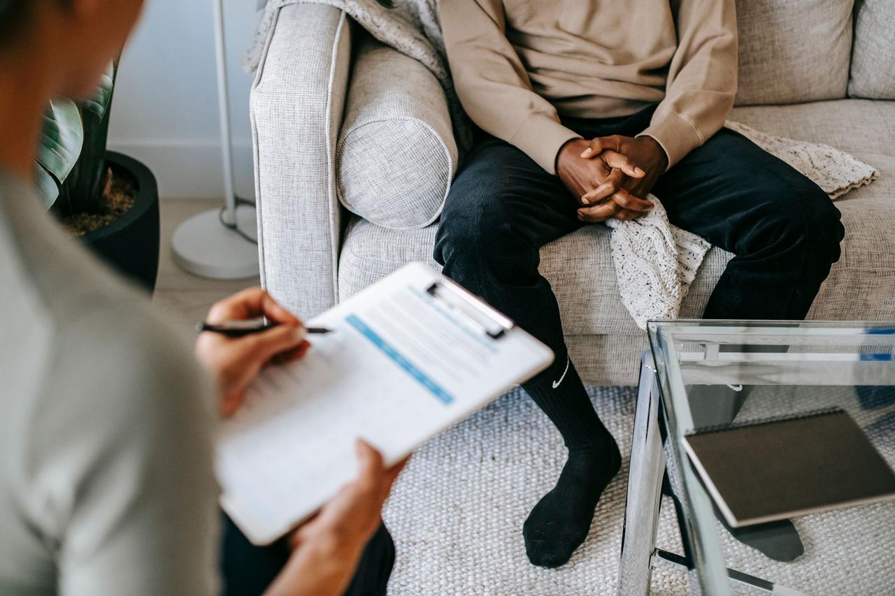 Blurred unrecognizable female therapist taking notes on clipboard while sitting in front of ethnic male patient with clasped hands during meeting