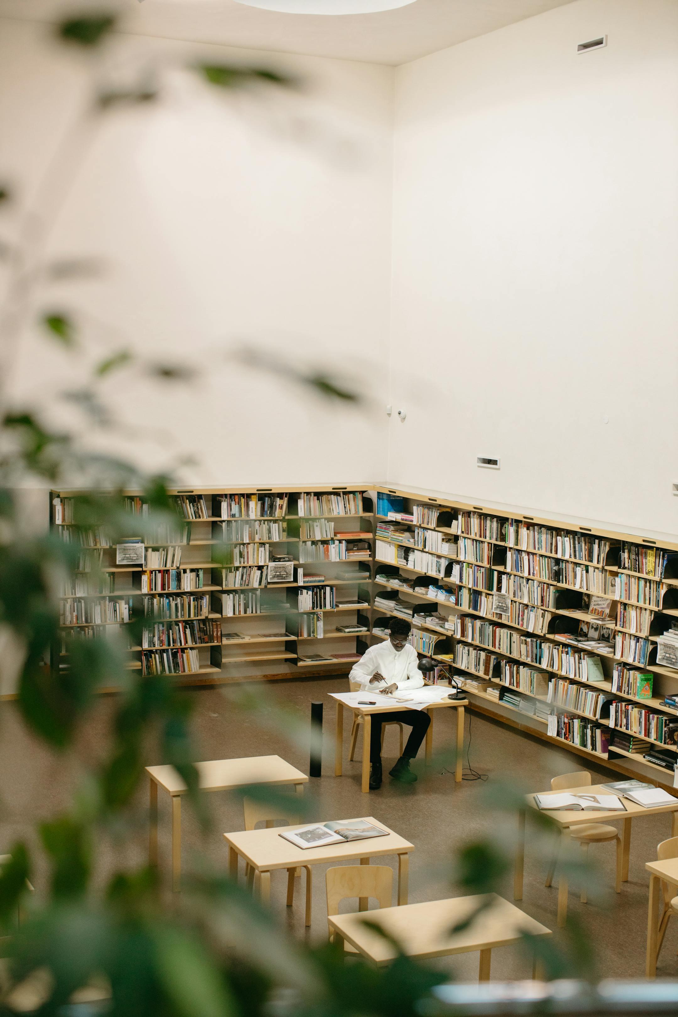 Adult man studying at a desk in a bright modern library, surrounded by bookshelves.