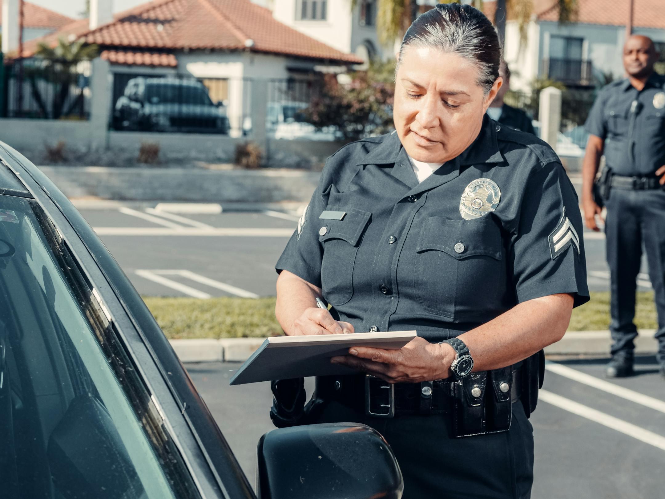 Police officer writing a ticket to a car in a sunny parking lot with a colleague in the background.