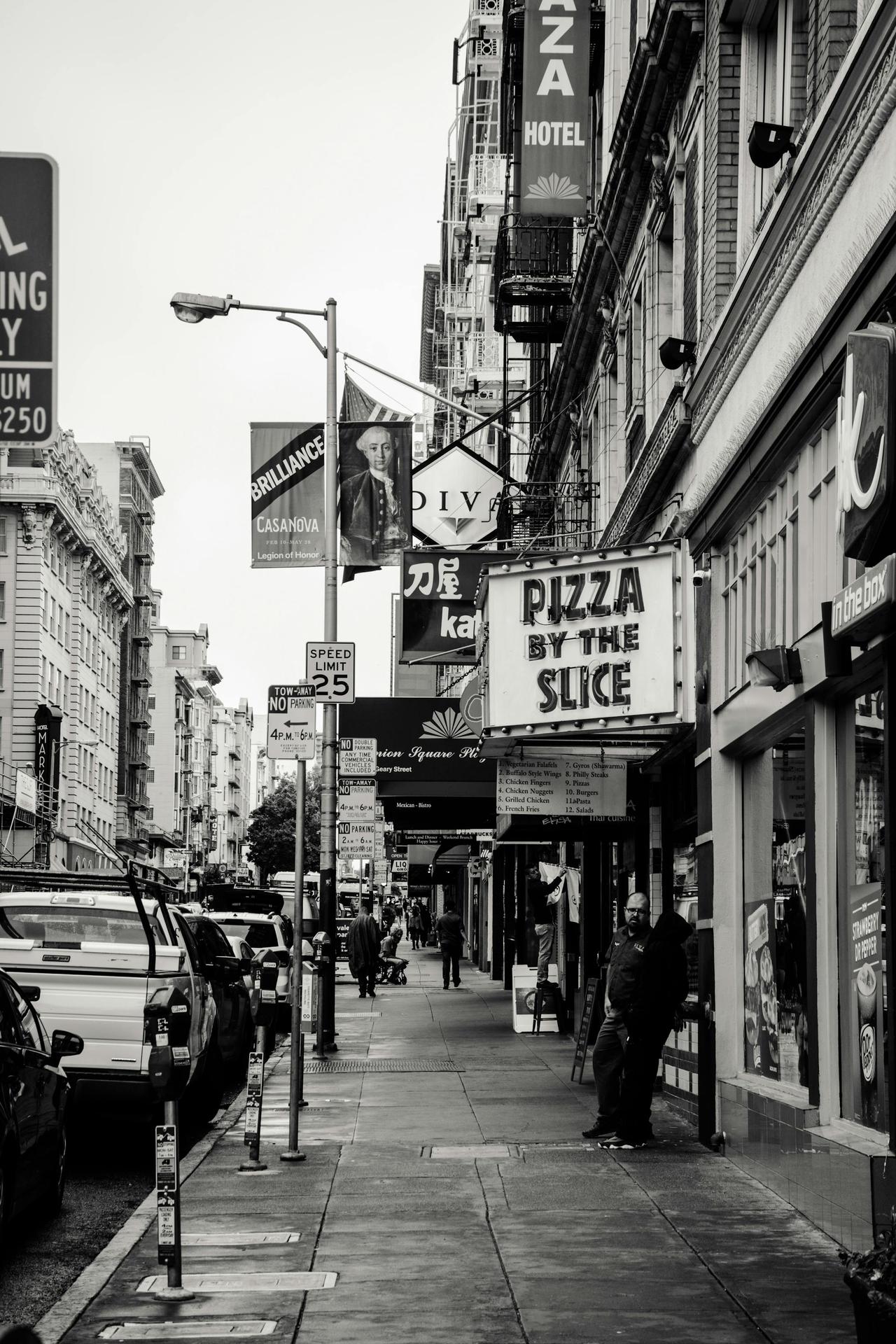 Black and white photo of a busy city street featuring storefronts, signs, and pedestrians.