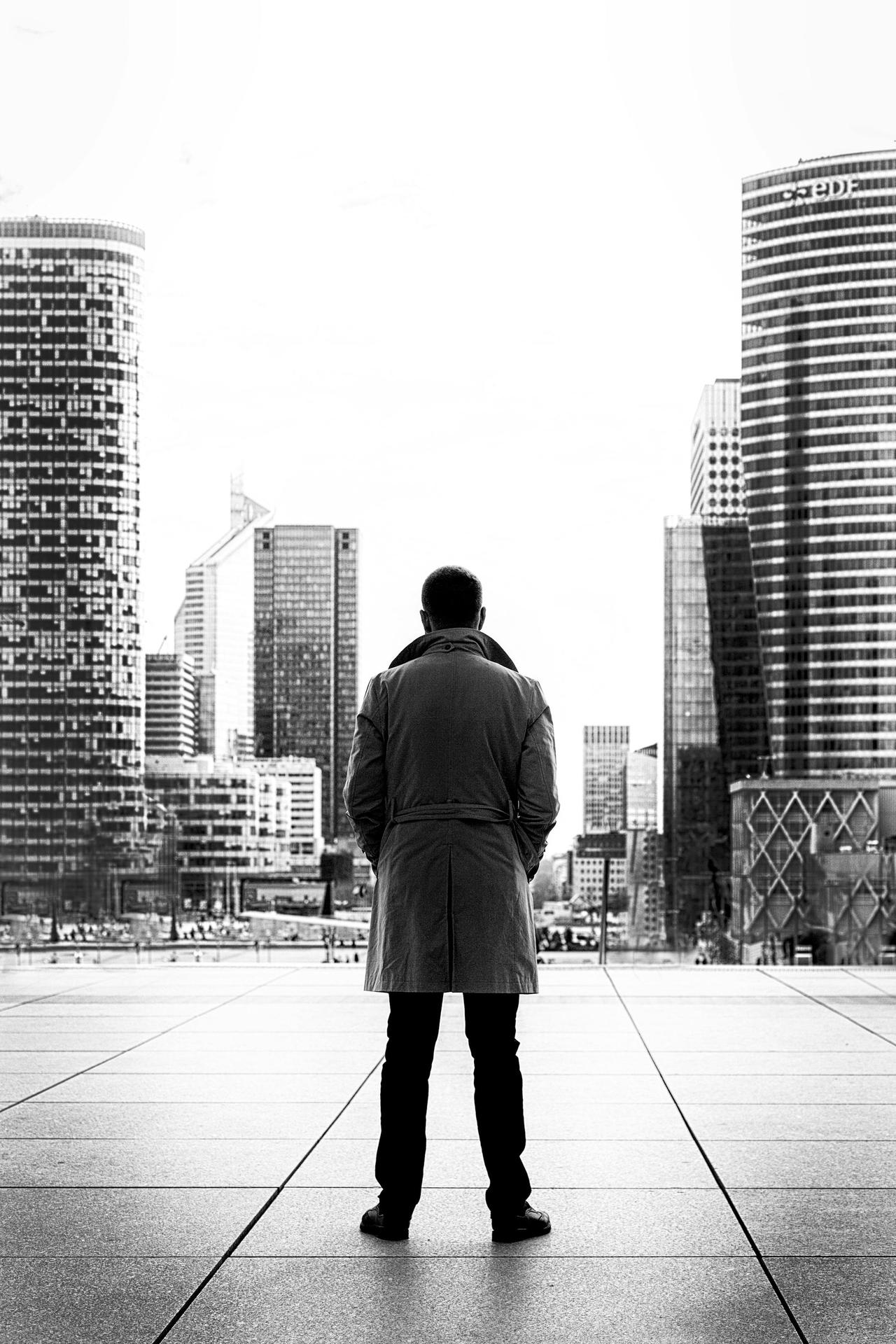 Silhouette of a man in a trench coat standing against a modern urban cityscape in black and white.