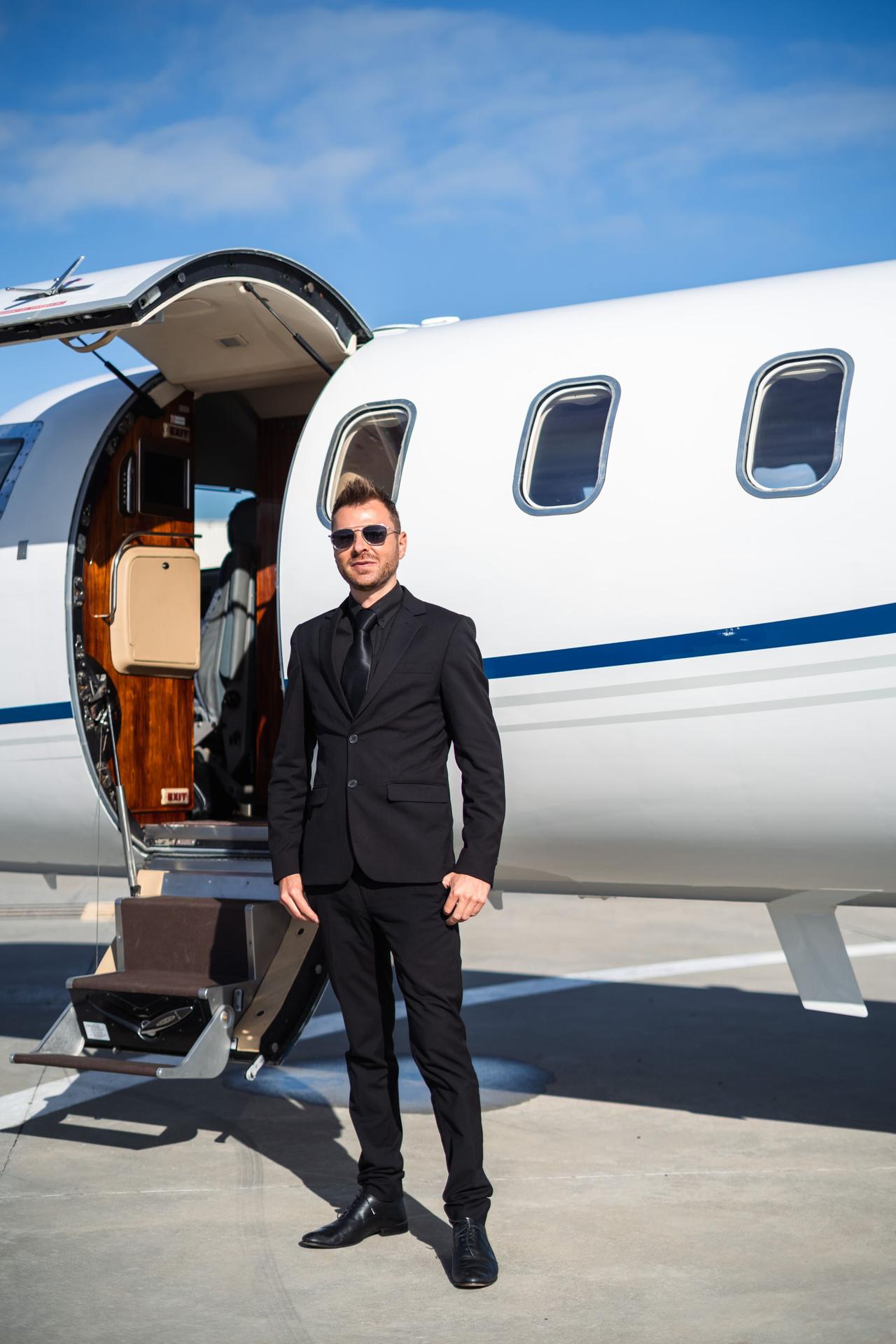 Stylish man in black suit poses confidently beside a private jet under clear blue skies.