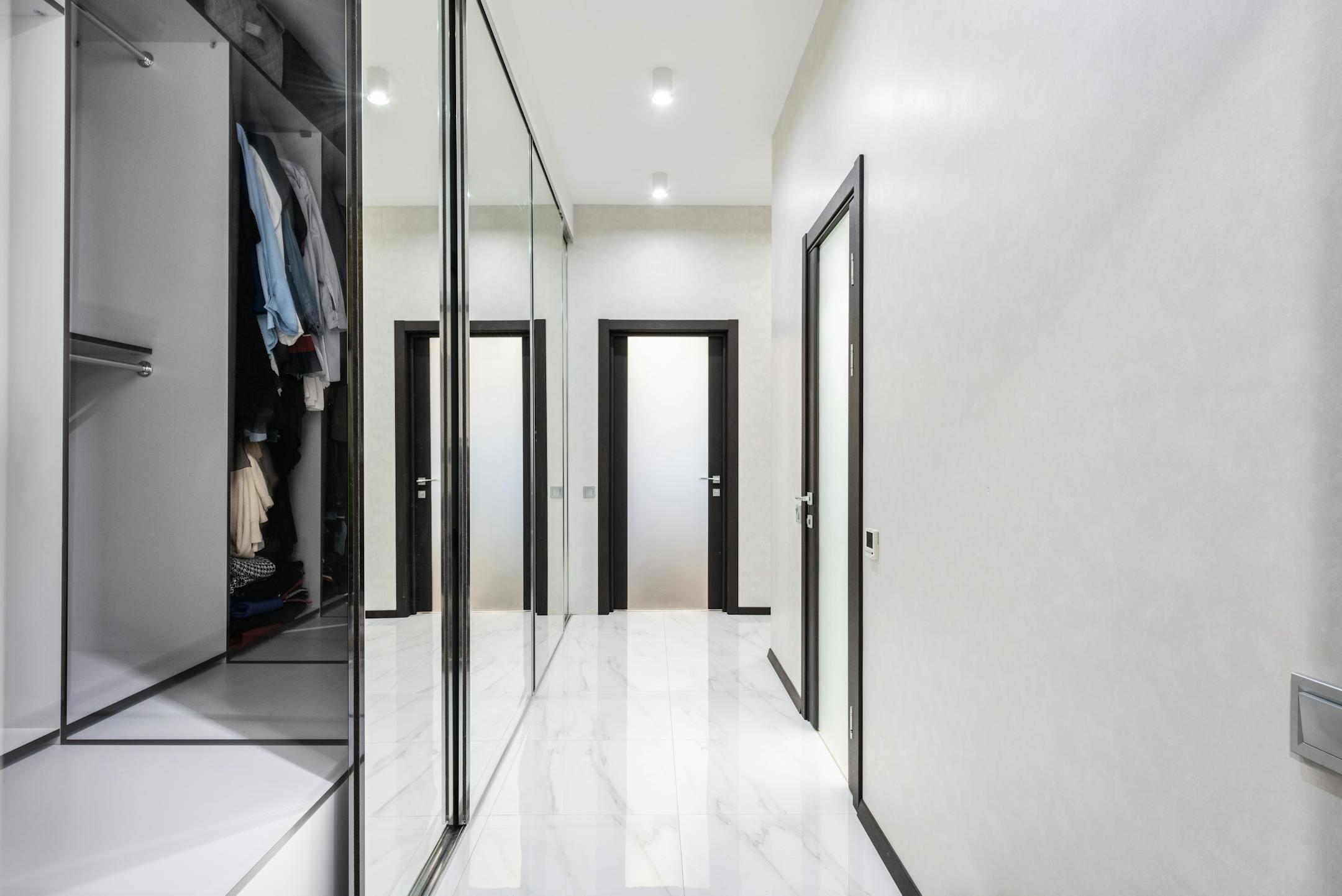Long light corridor with white walls and opened wardrobe with cloth in modern apartment with closed doors and glowing lamps