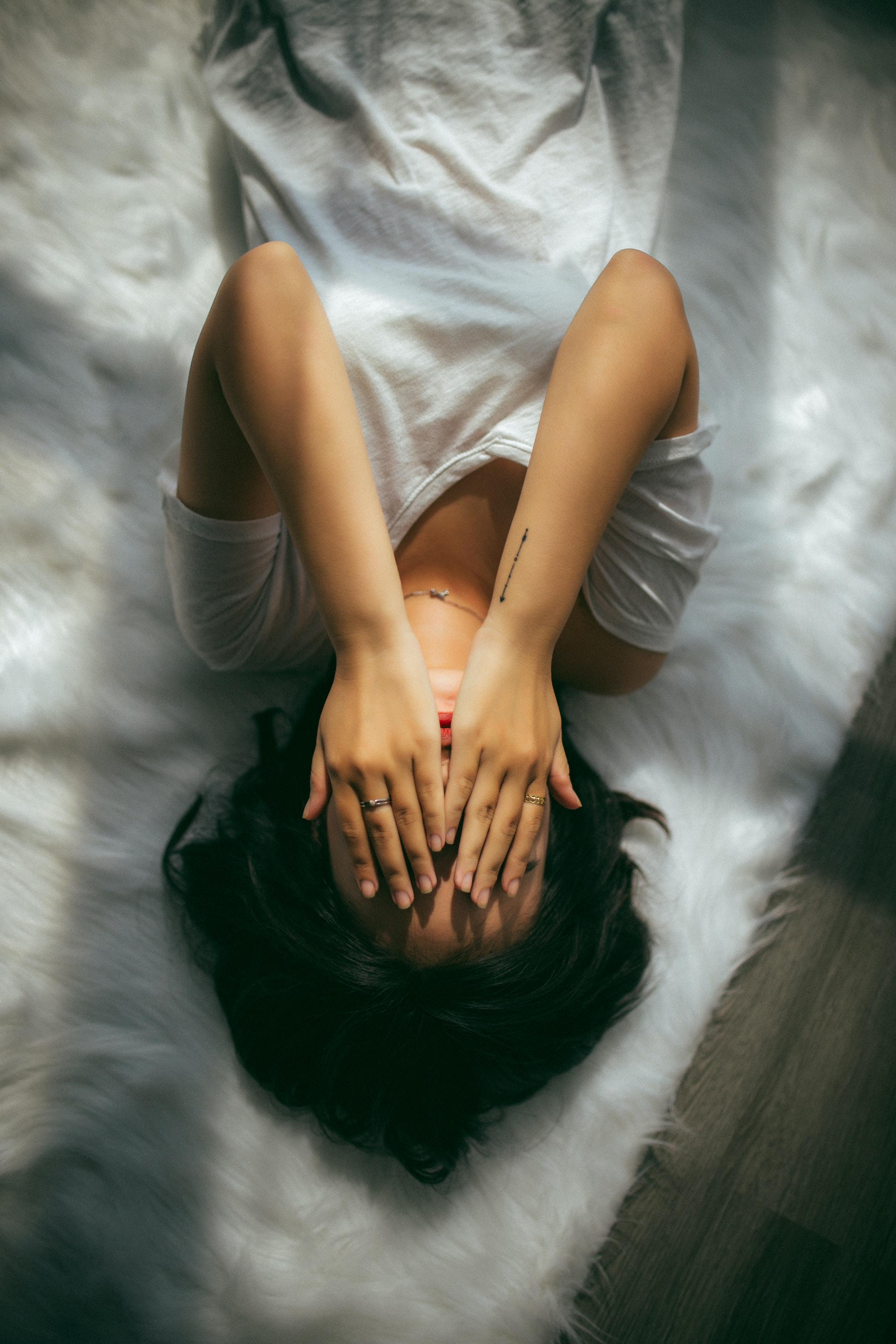 a woman lays down on a white bed, upside down to the view, with her hands covering her face in distress.