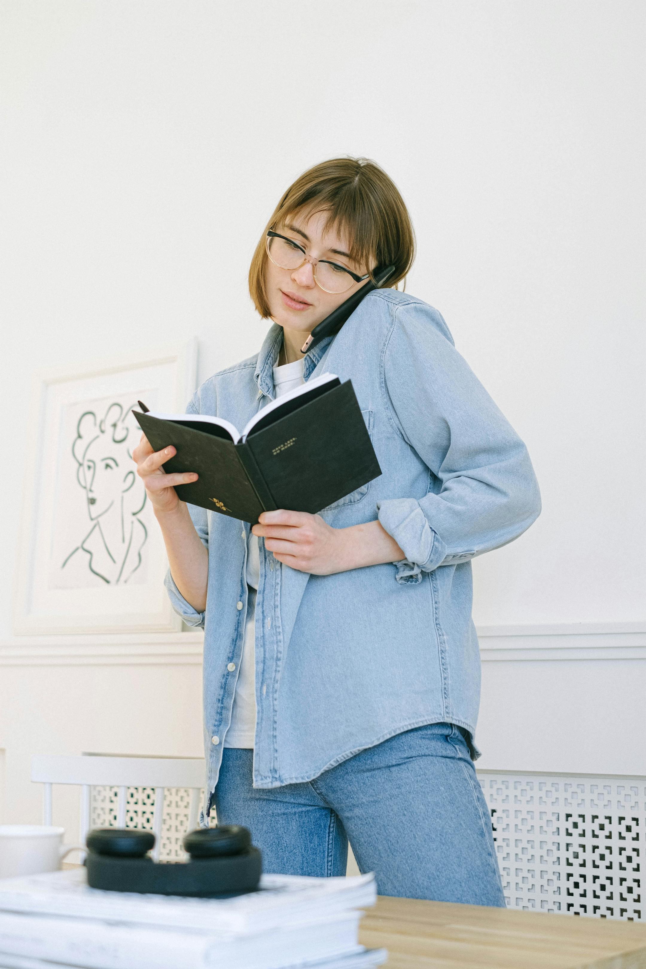 Young woman multitasking in a home office, holding an agenda and making a phone call.