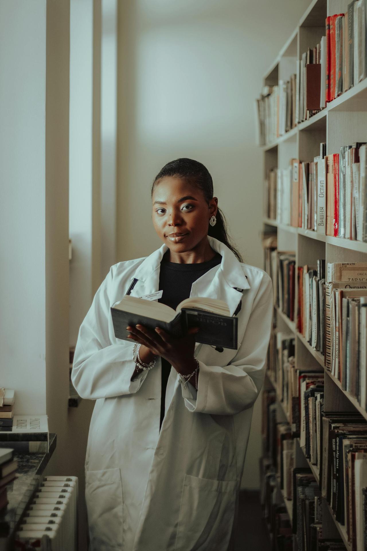 A focused woman in a lab coat reading a book in a library, showcasing education and learning.