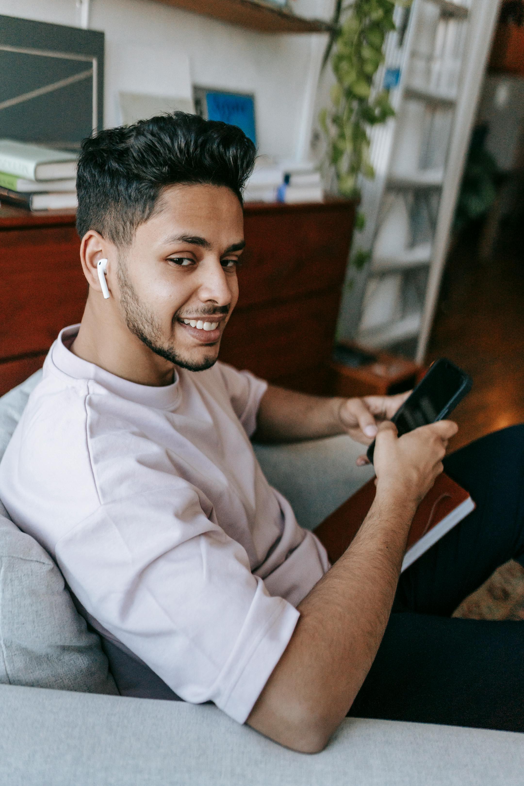 Side view happy young male using mobile phone and listening to music in earbuds while resting in cozy armchair