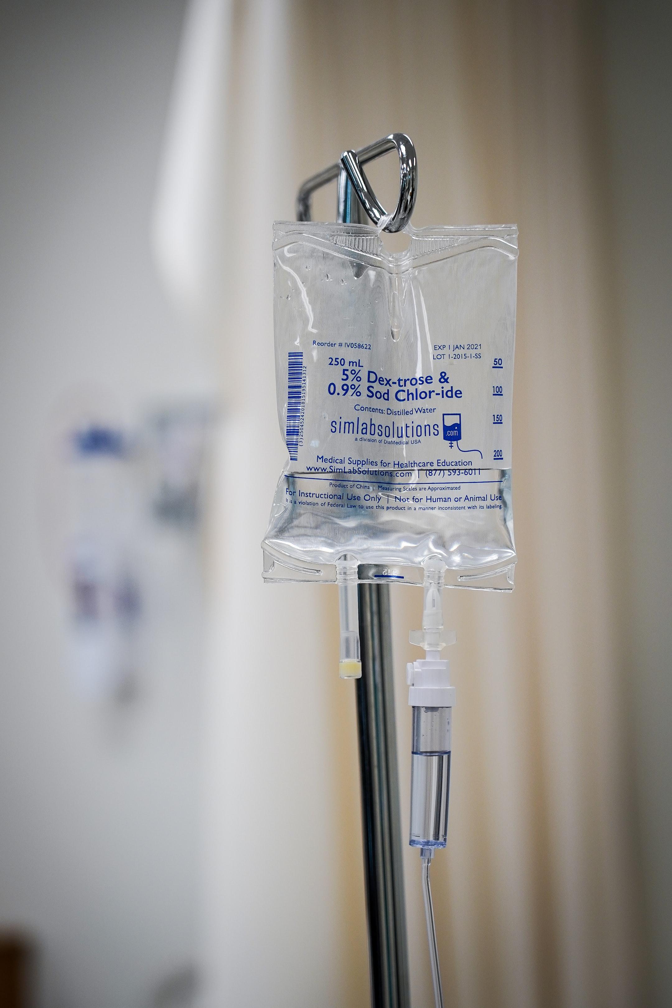 An IV bag hangs from a pole in a hospital room. A tube runs from the bag to a patient's arm. 