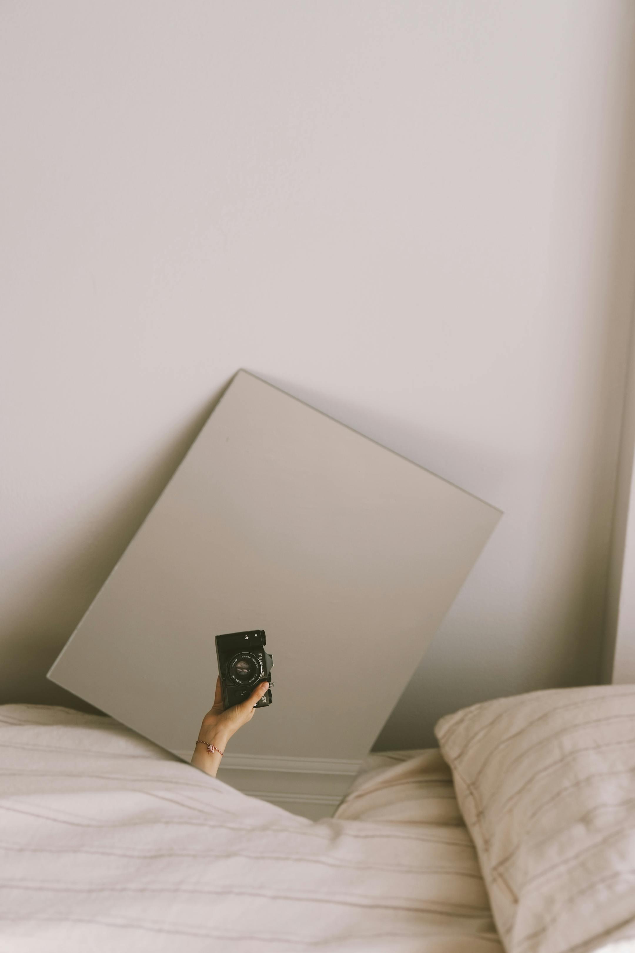 A hand holding a camera reflected in a mirror on a bed, capturing a creative indoor scene.