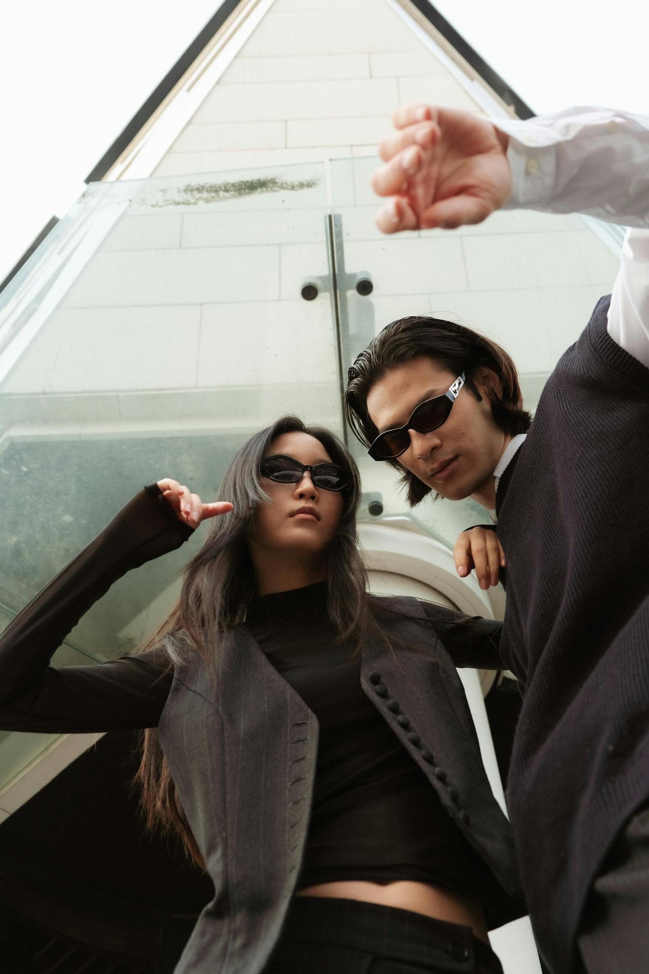 Stylish man and woman in black outfits with sunglasses posing with architectural backdrop.