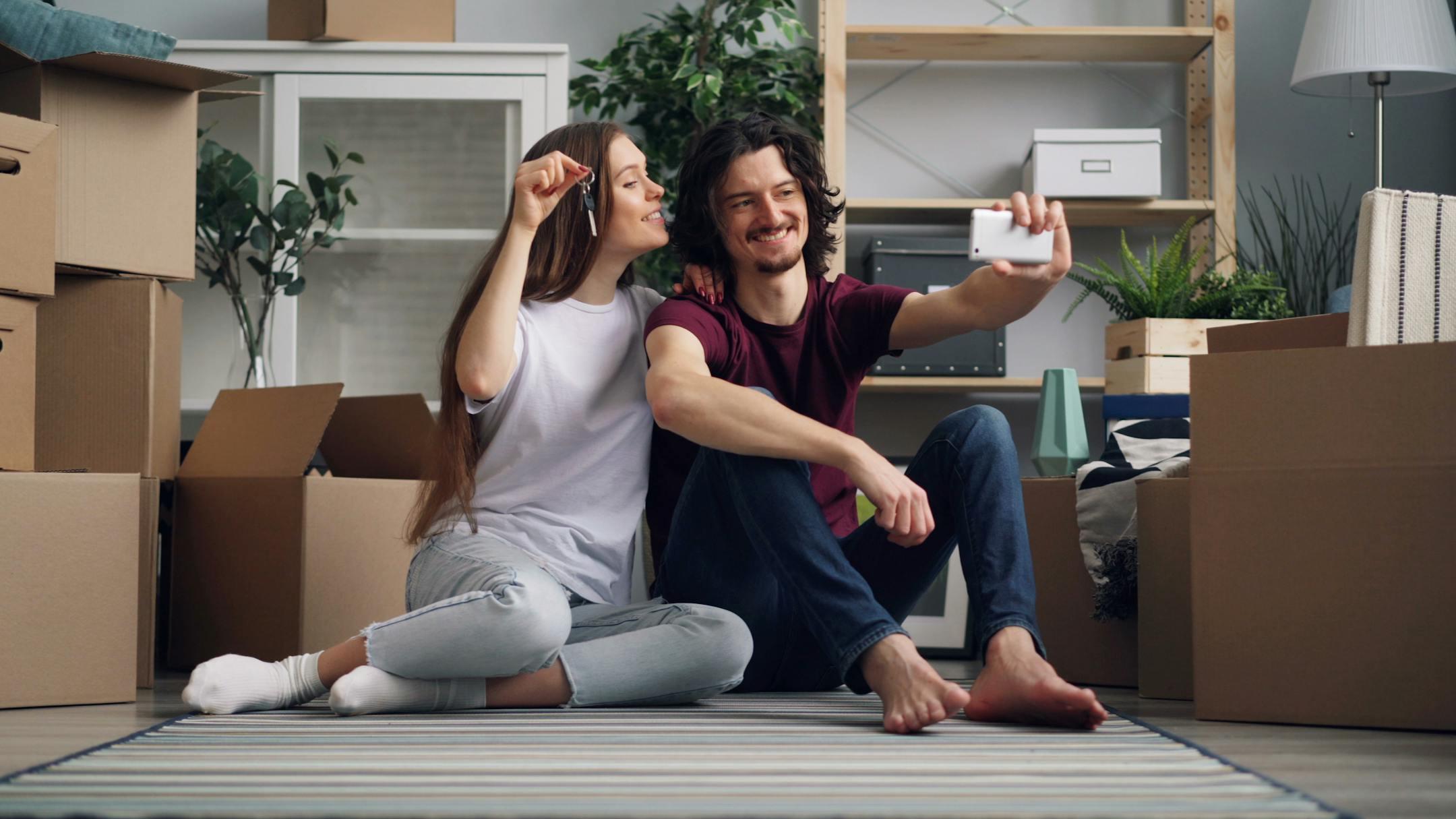 Smiling couple taking a selfie while moving into their new home, surrounded by boxes.