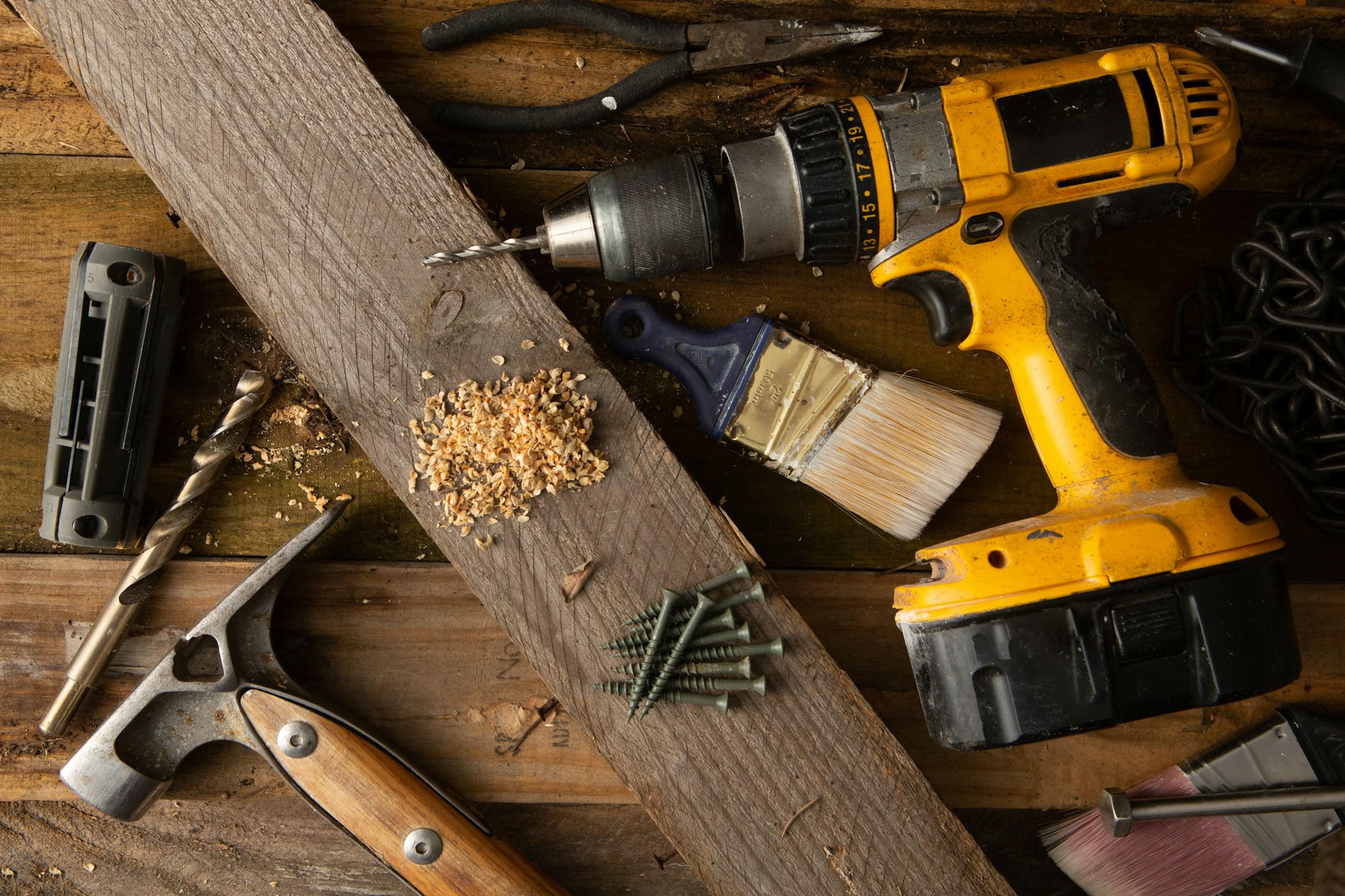 A variety of tools, including a hammer and cordless drill, sit on top of a table made of wood boards. 