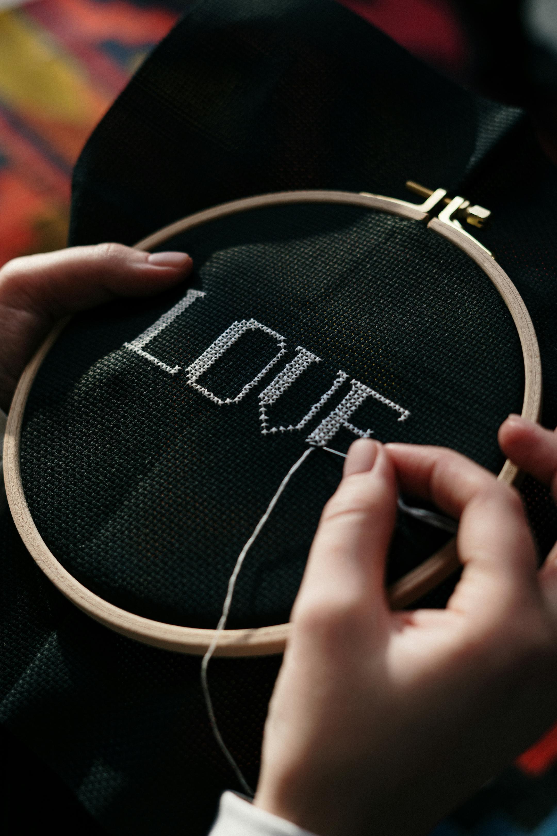 Close-up of hands embroidering 'Love' on black fabric with hoop.