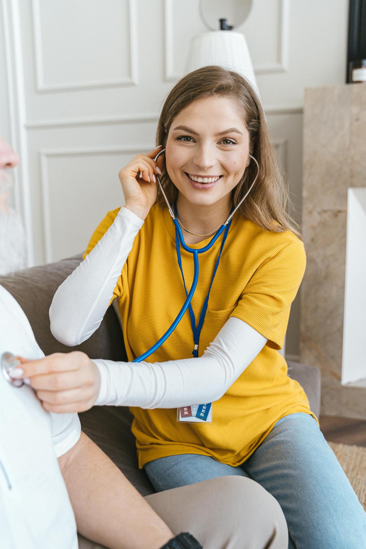 A volunteer nurse with a stethoscope smiles while assisting an elderly patient indoors.