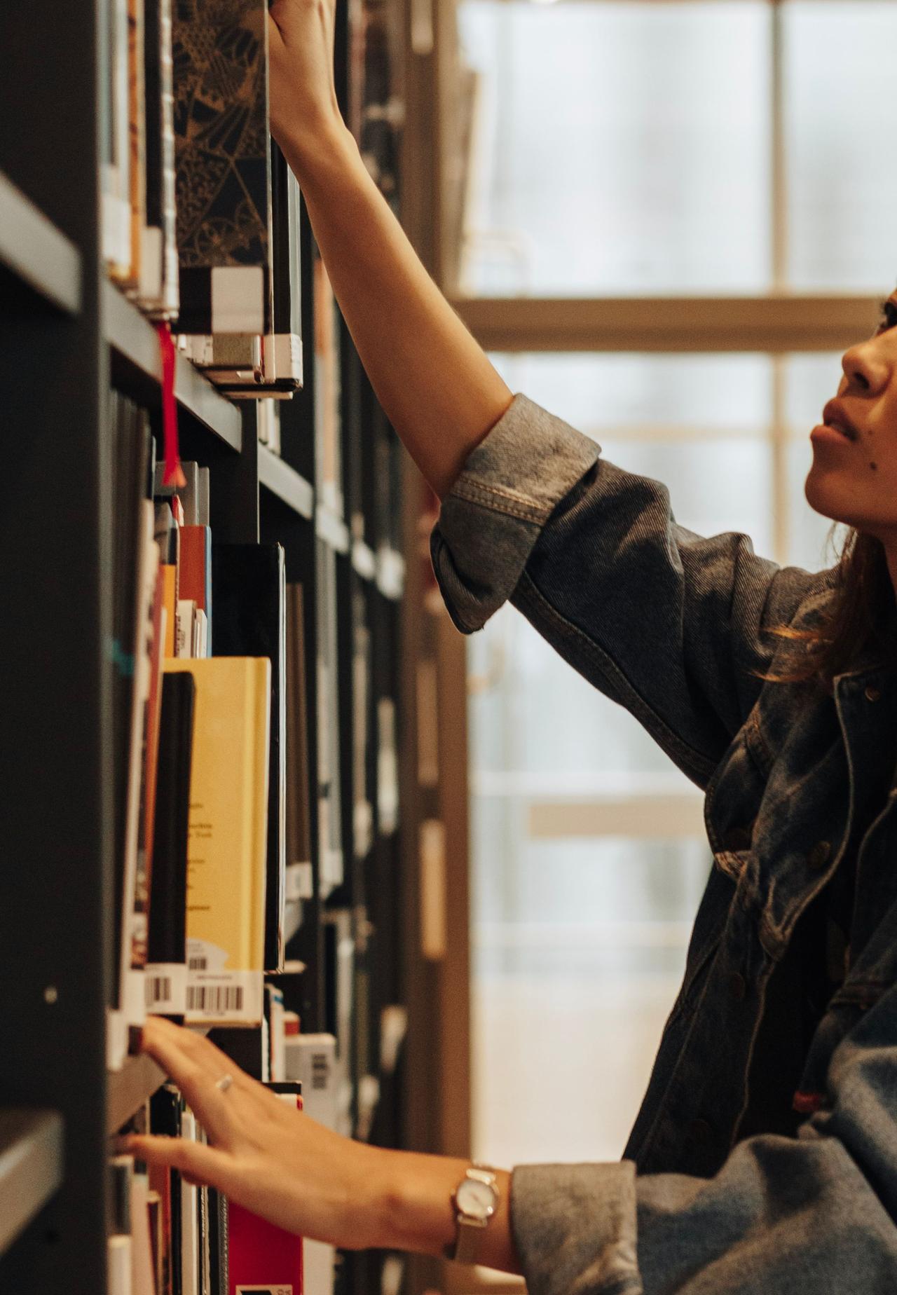 A woman reaching for a book in a well-lit modern library aisle.