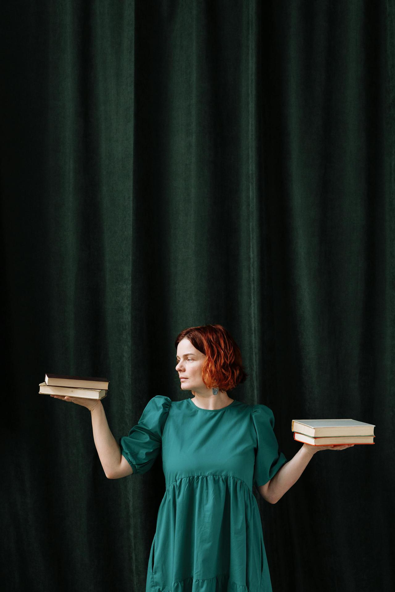 Woman in green dress balancing books with dark curtain backdrop.
