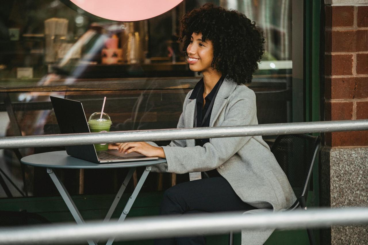 Cheerful businesswoman with afro hairstyle working on laptop outdoors at café.