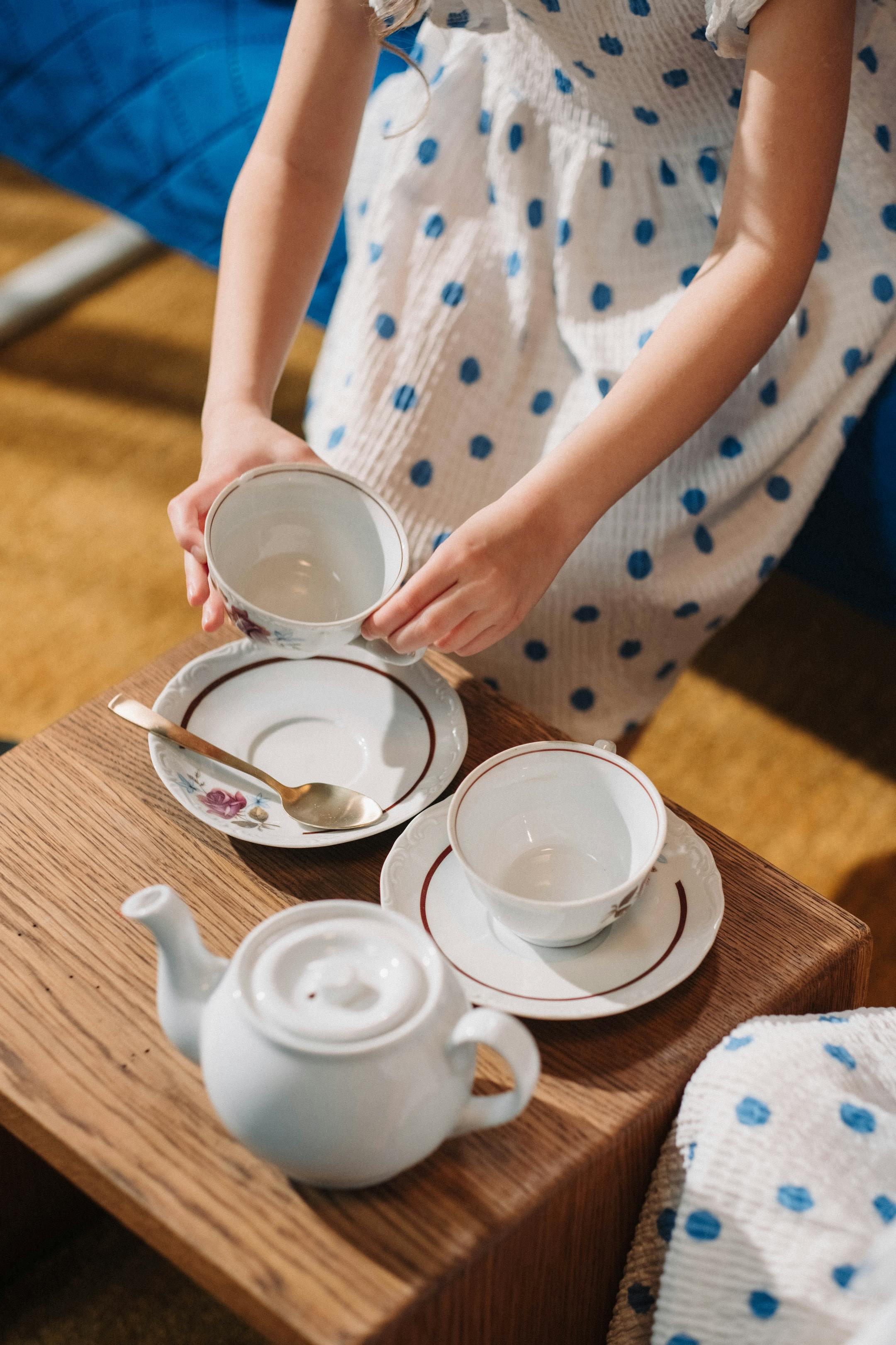 Young girl arranging delicate porcelain tea cups on a wooden table during a playful tea party indoors.