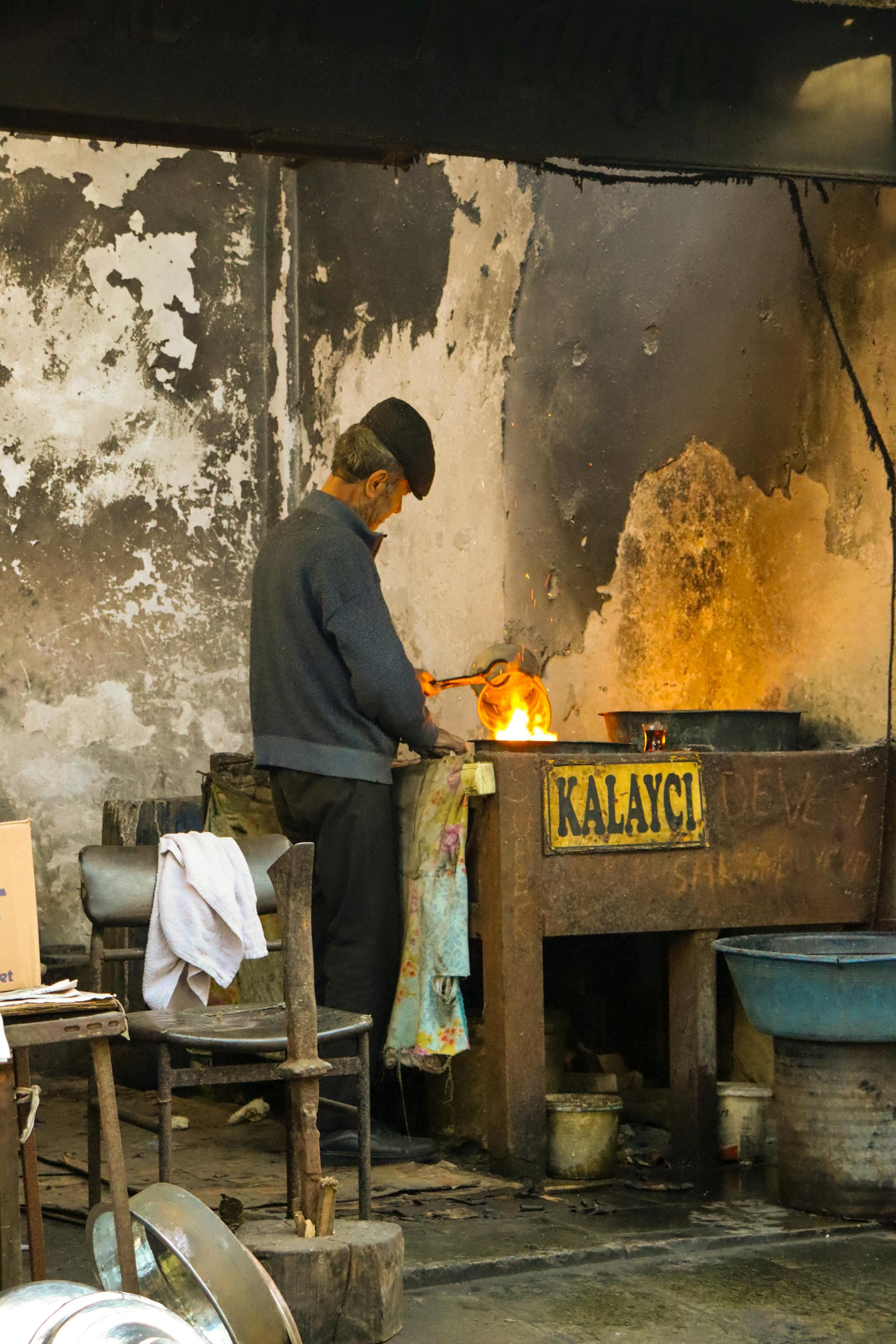 Old craftsman working with fire in a rustic workshop. Sign reads 'Kalayci'.