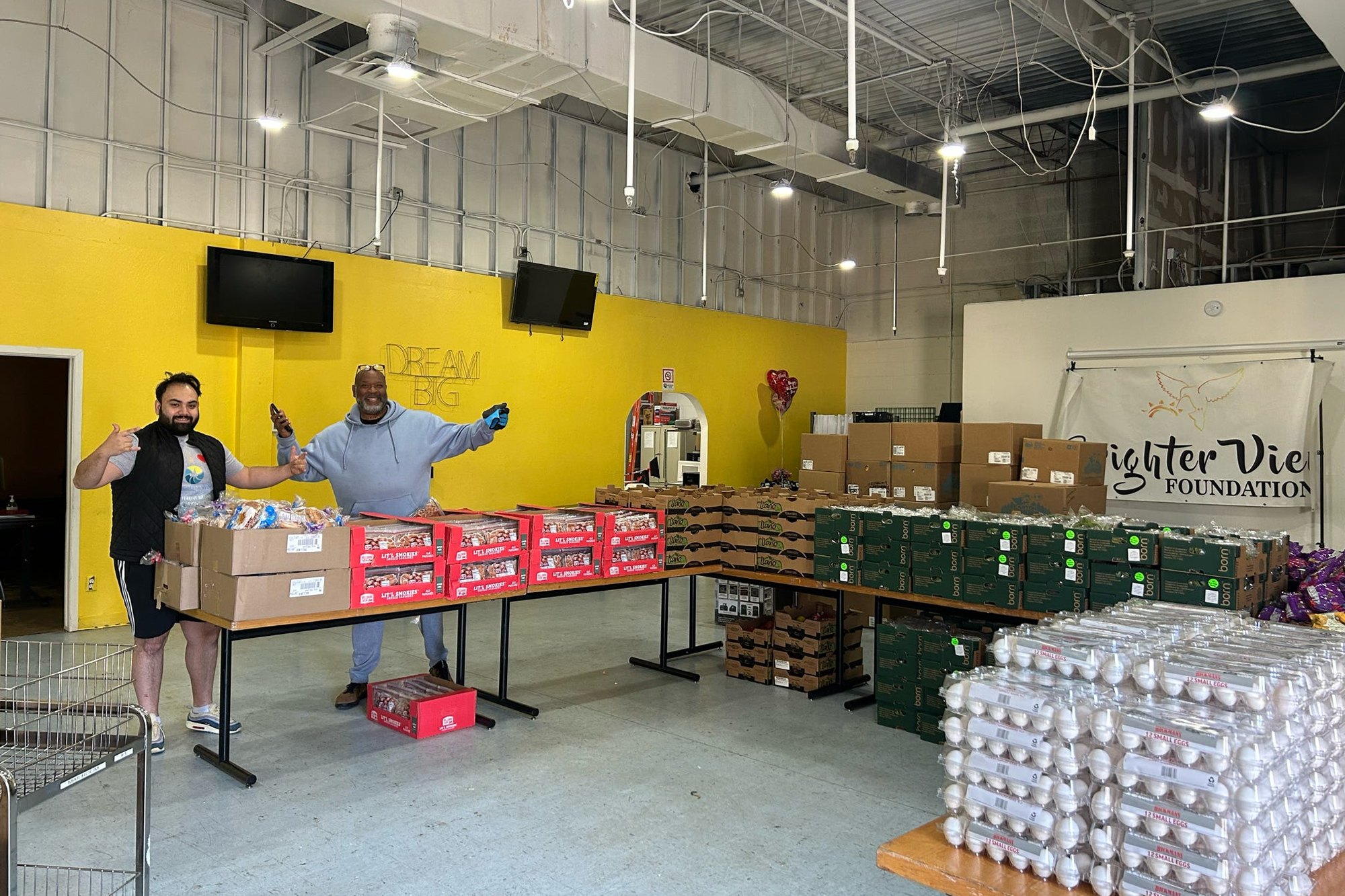 Two staff members welcoming visitors in front of fully stocked pantry tables.
