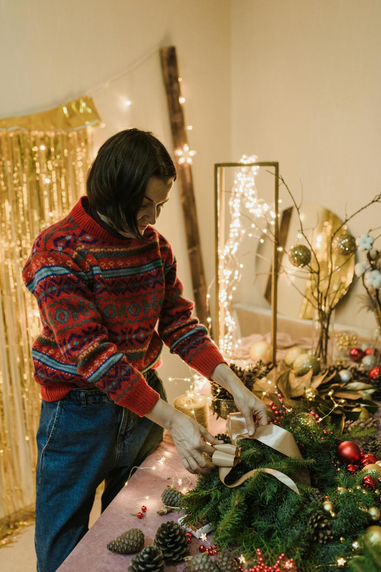 Woman in a festive sweater crafting a Christmas wreath indoors decorated with lights.