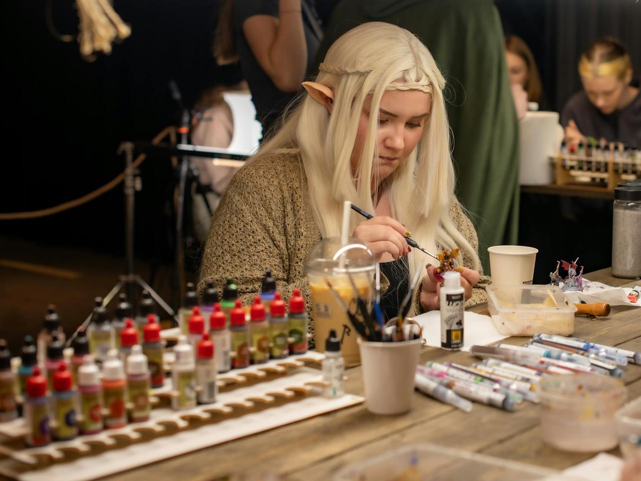 A woman cosplays as an elf while painting miniatures at an indoor convention.