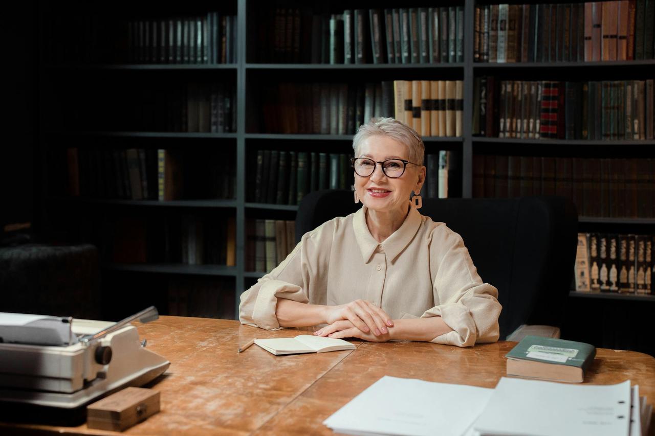 Senior woman sitting at desk in library, smiling at camera, with books and typewriter.