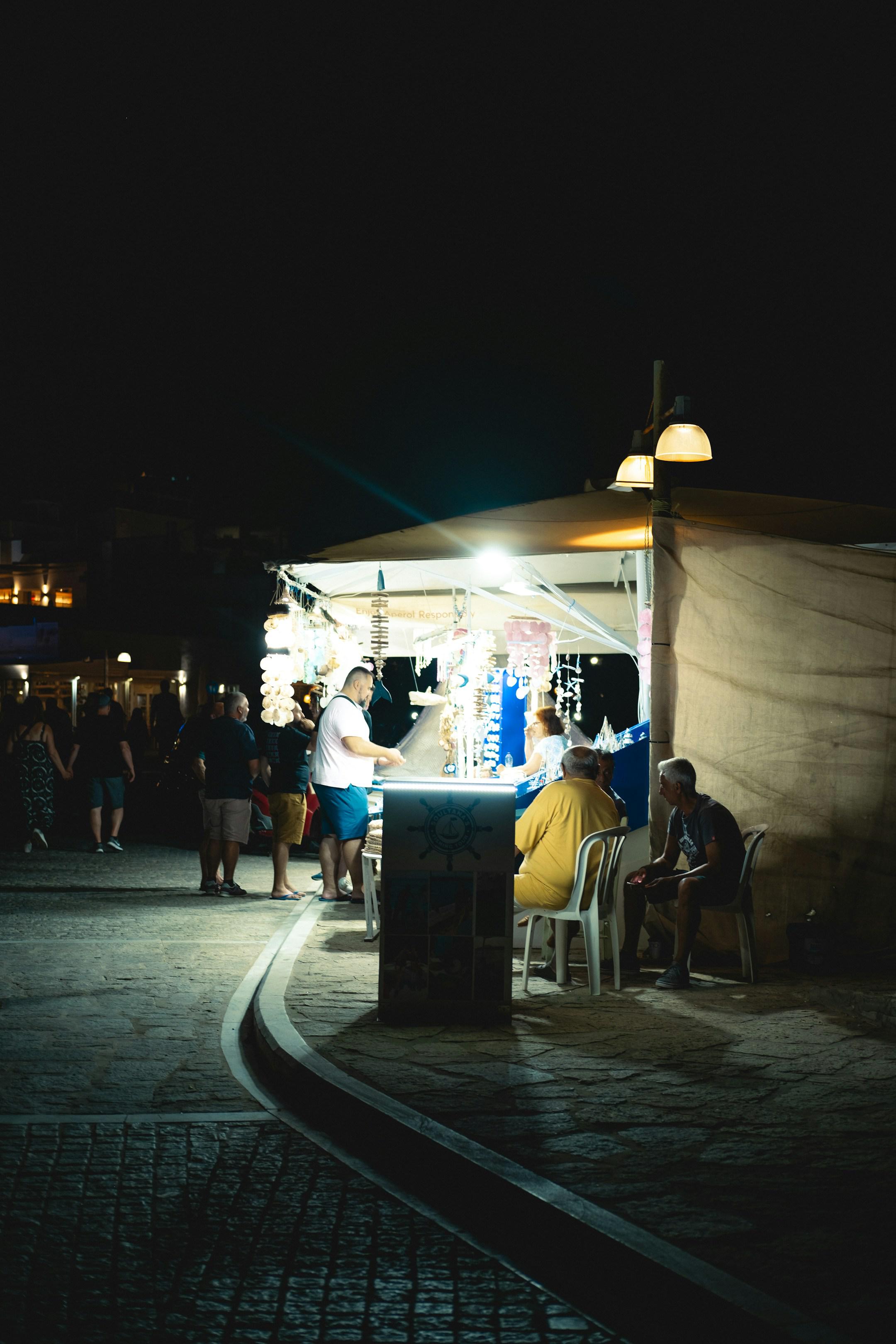 A vibrant night market scene with a well-lit stall selling trinkets and jewelry. People are gathered around, browsing and interacting with the vendor. The background features dimly lit buildings, adding depth to the lively atmosphere. The image captures the essence of a bustling night market filled with activity and charm.