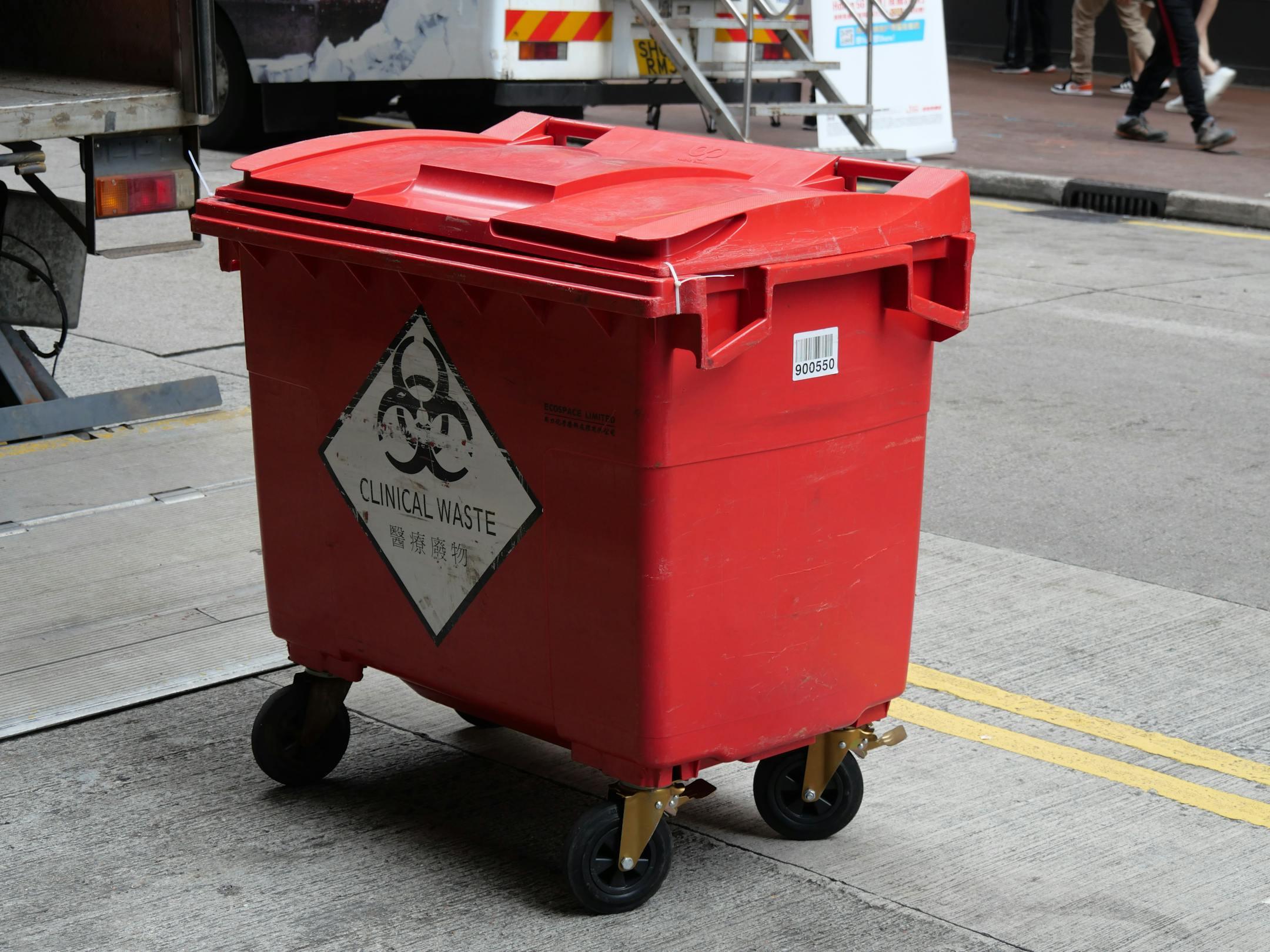 A red clinical waste bin labeled with biohazard symbol on a city street indicating proper waste management.