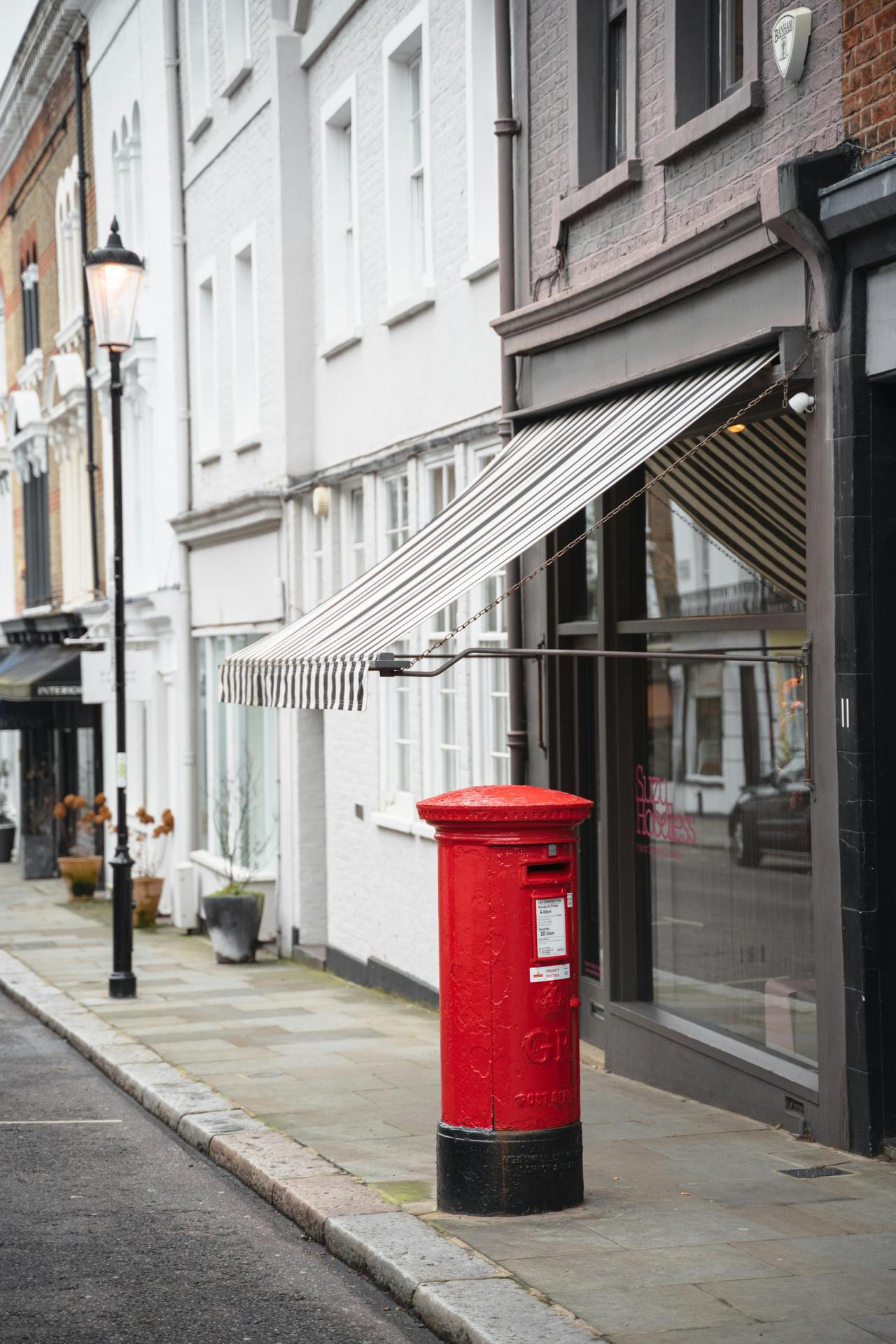 Red mail post box for sending letters located on sidewalk near road and aged residential house with canopy in city