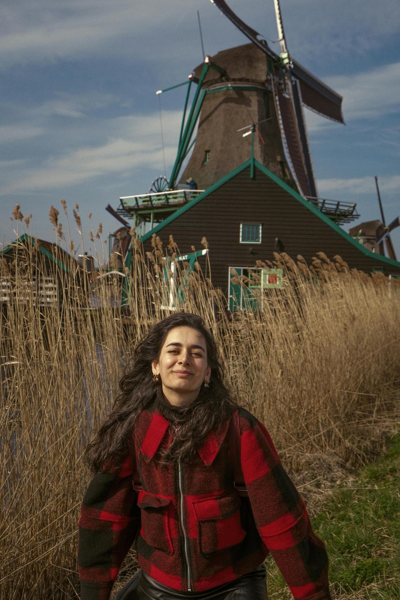 A woman smiles joyfully near an iconic Dutch windmill amidst tall reeds under a clear blue sky.