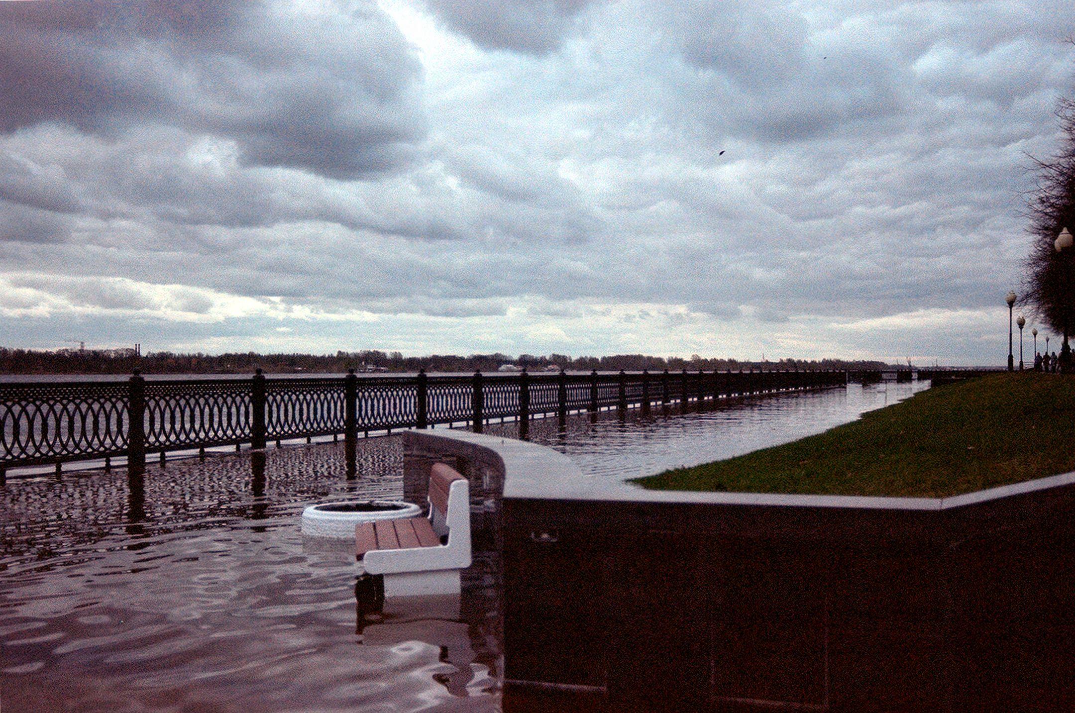 A flooded walkway by the river with overcast skies and empty benches submerged.