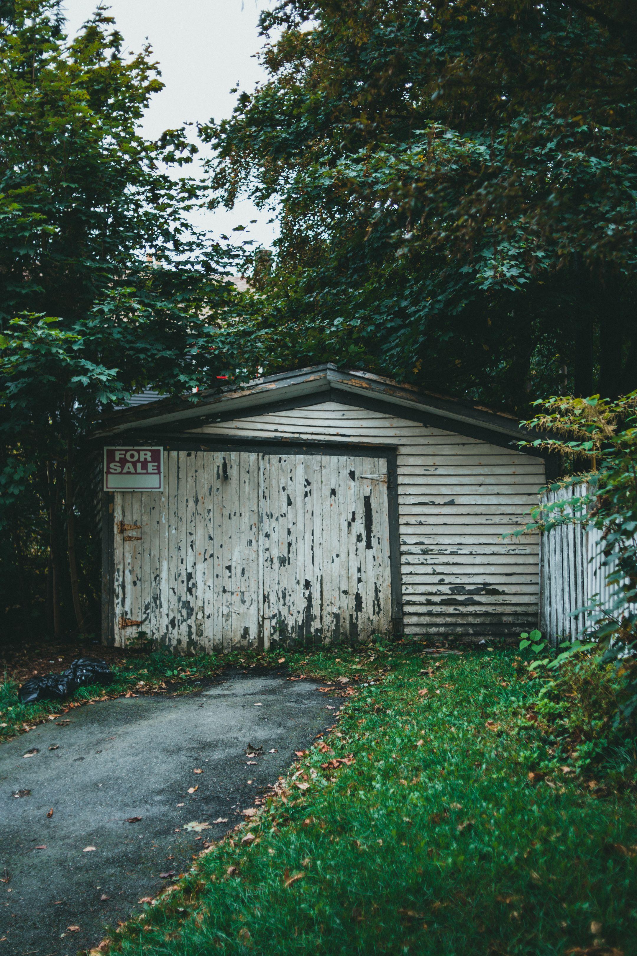 Old wooden garage with 'For Sale' sign surrounded by overgrown greenery.