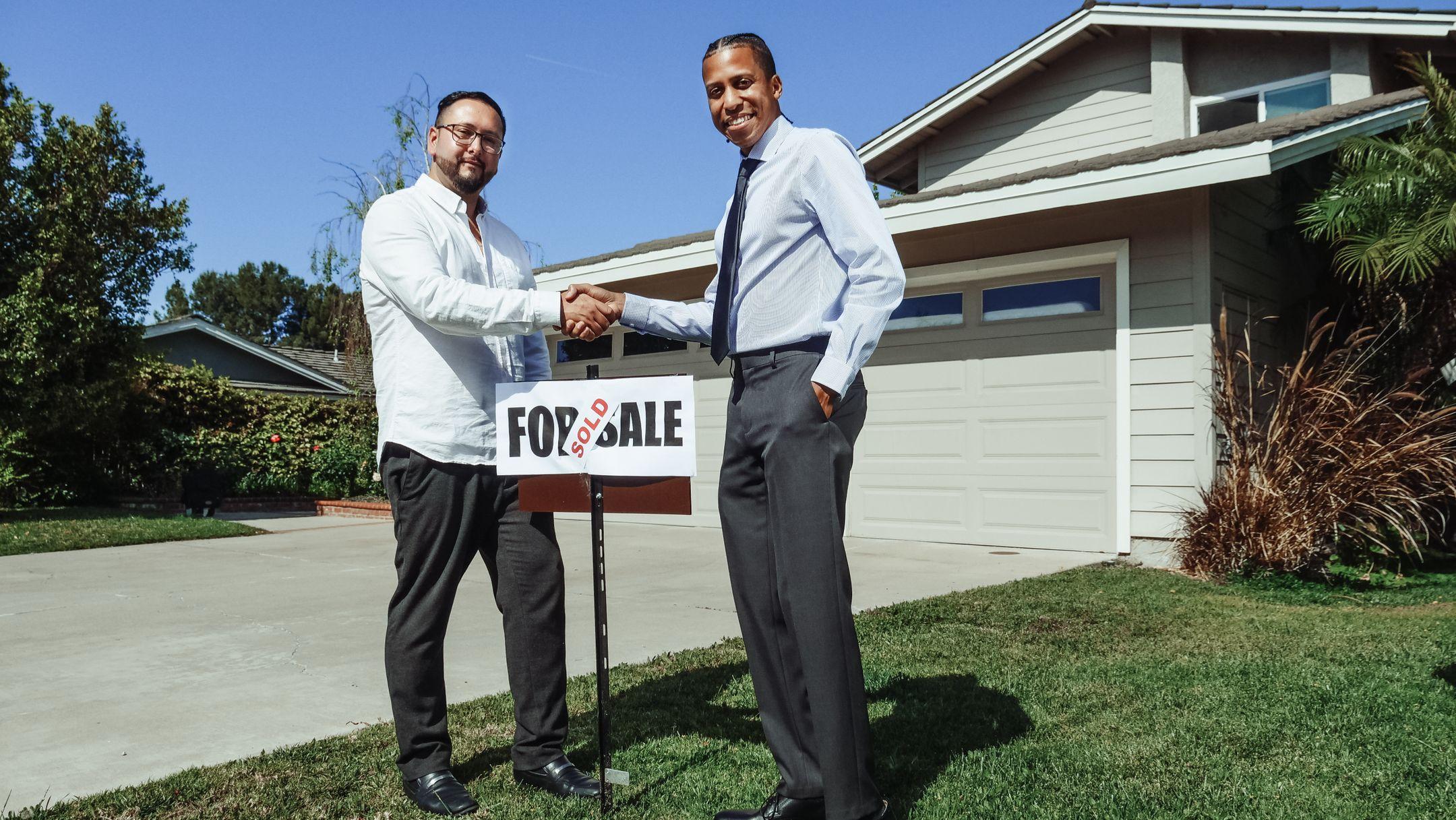 Two men shake hands in front of a new home with a sold sign, symbolizing a successful real estate deal.