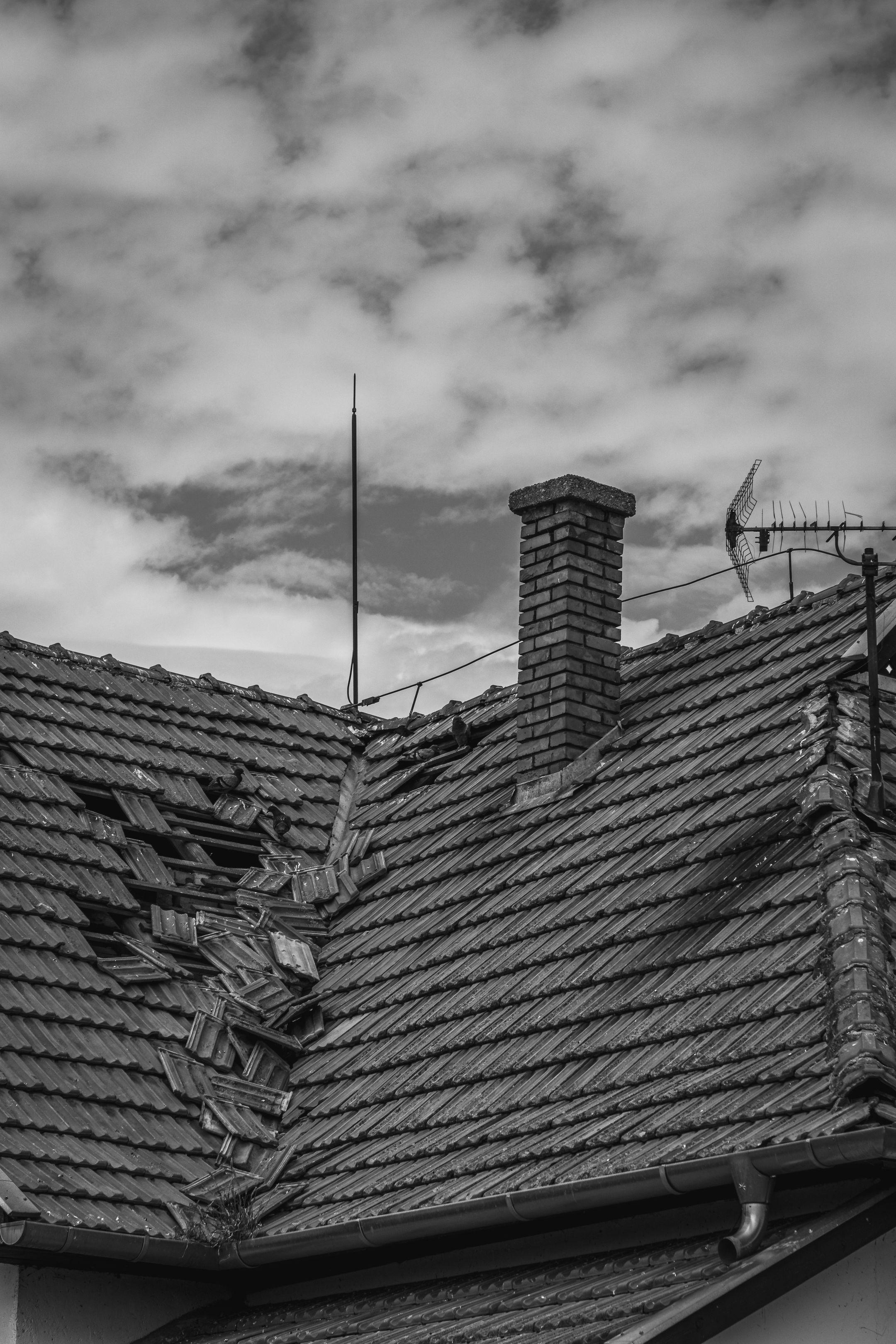 A grayscale photo of a broken roof with a chimney and antenna under a cloudy sky.