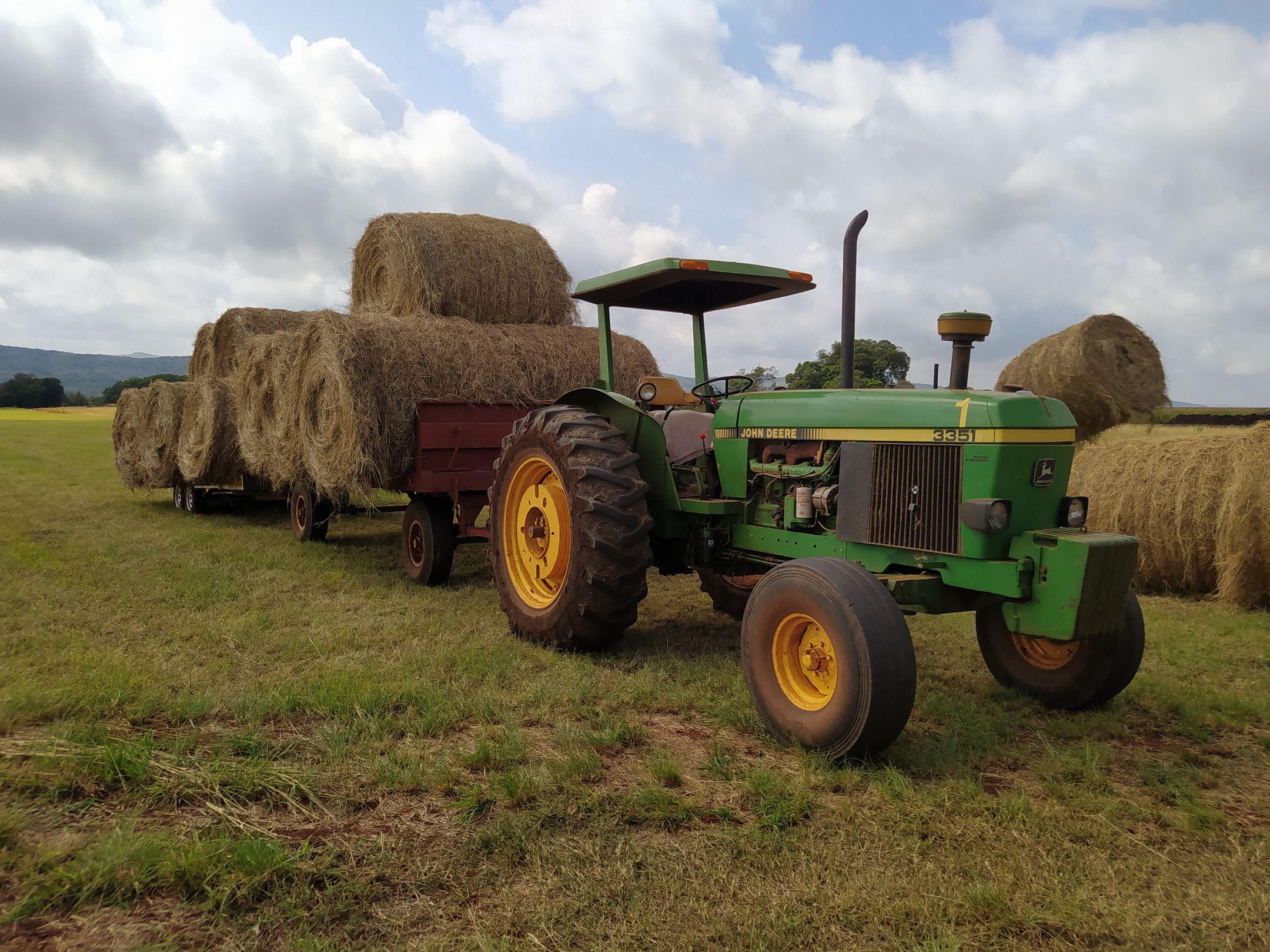 A green tractor pulling a trailer loaded with large hay bales in a rural field.